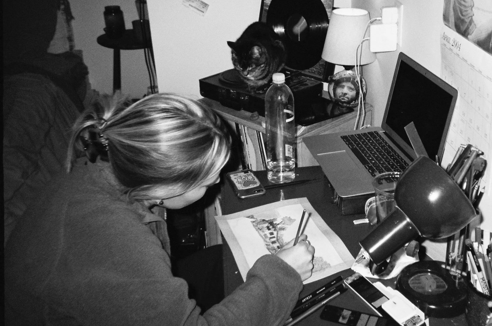 A woman seated at a cluttered desk, drawing a painting of a house with trees and mountains, with a water bottle, a cell phone, pens, and a lamp on the desk. There is a laptop and a stereo with a cat on top in the background.