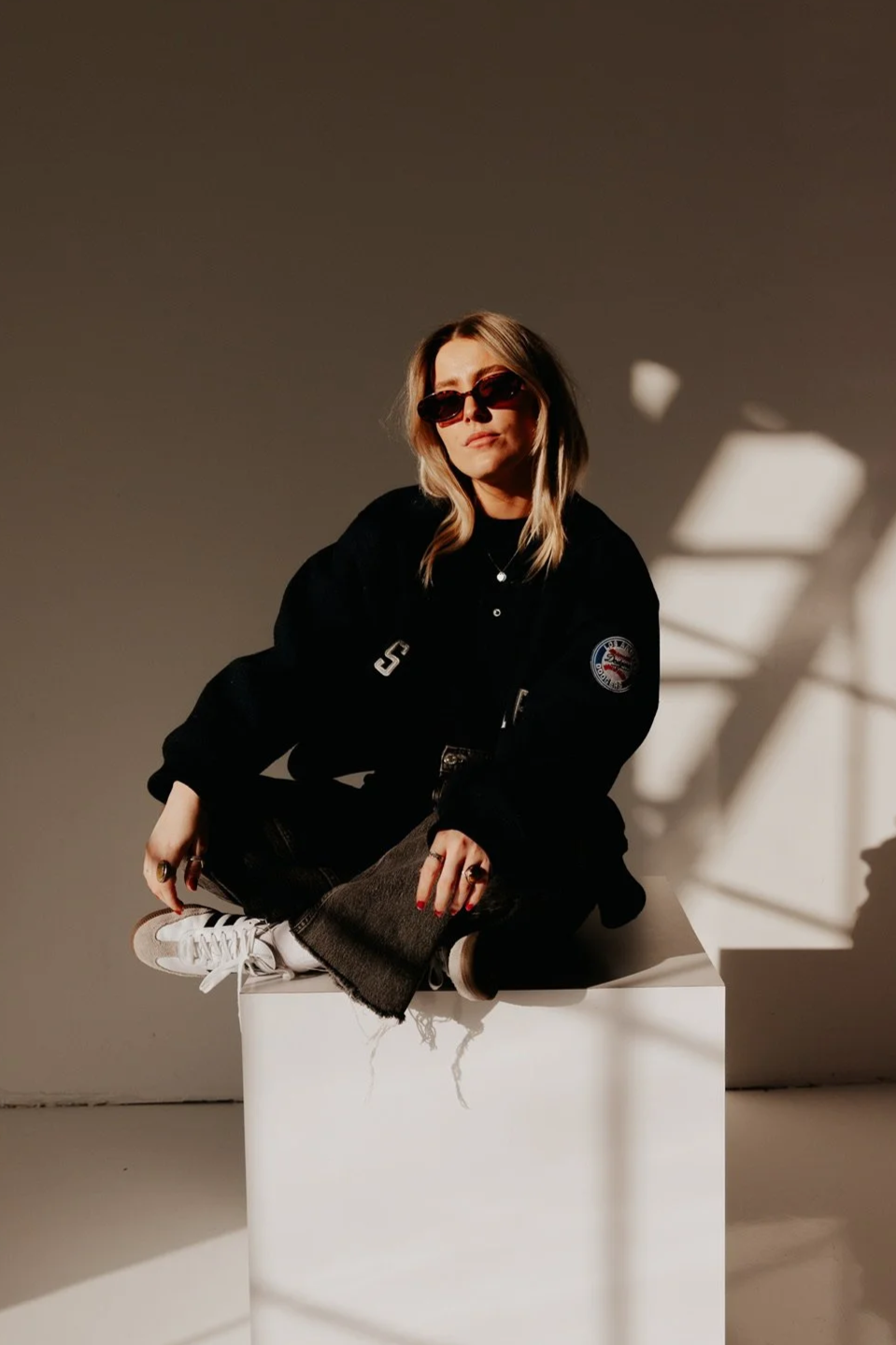A woman with blonde hair wearing sunglasses, a black jacket, and sneakers, sitting cross-legged on a white cube, with shadows cast on the beige wall behind her.