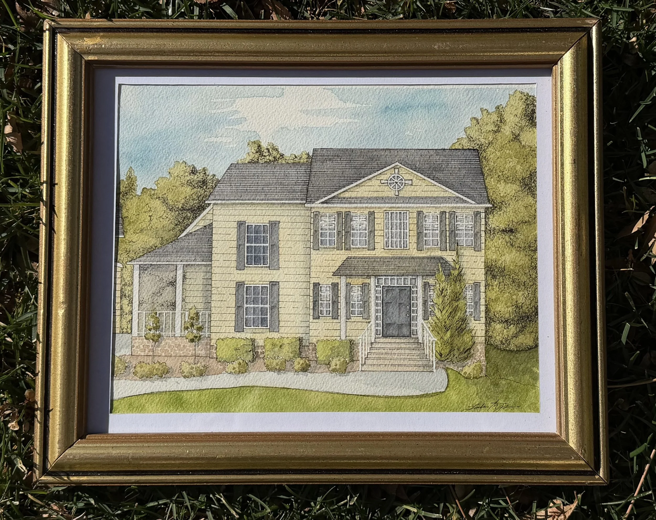 Colorful watercolor painting of a yellow house with gray shutters, surrounded by green trees and bushes, under a partly cloudy sky, framed in a gold frame.