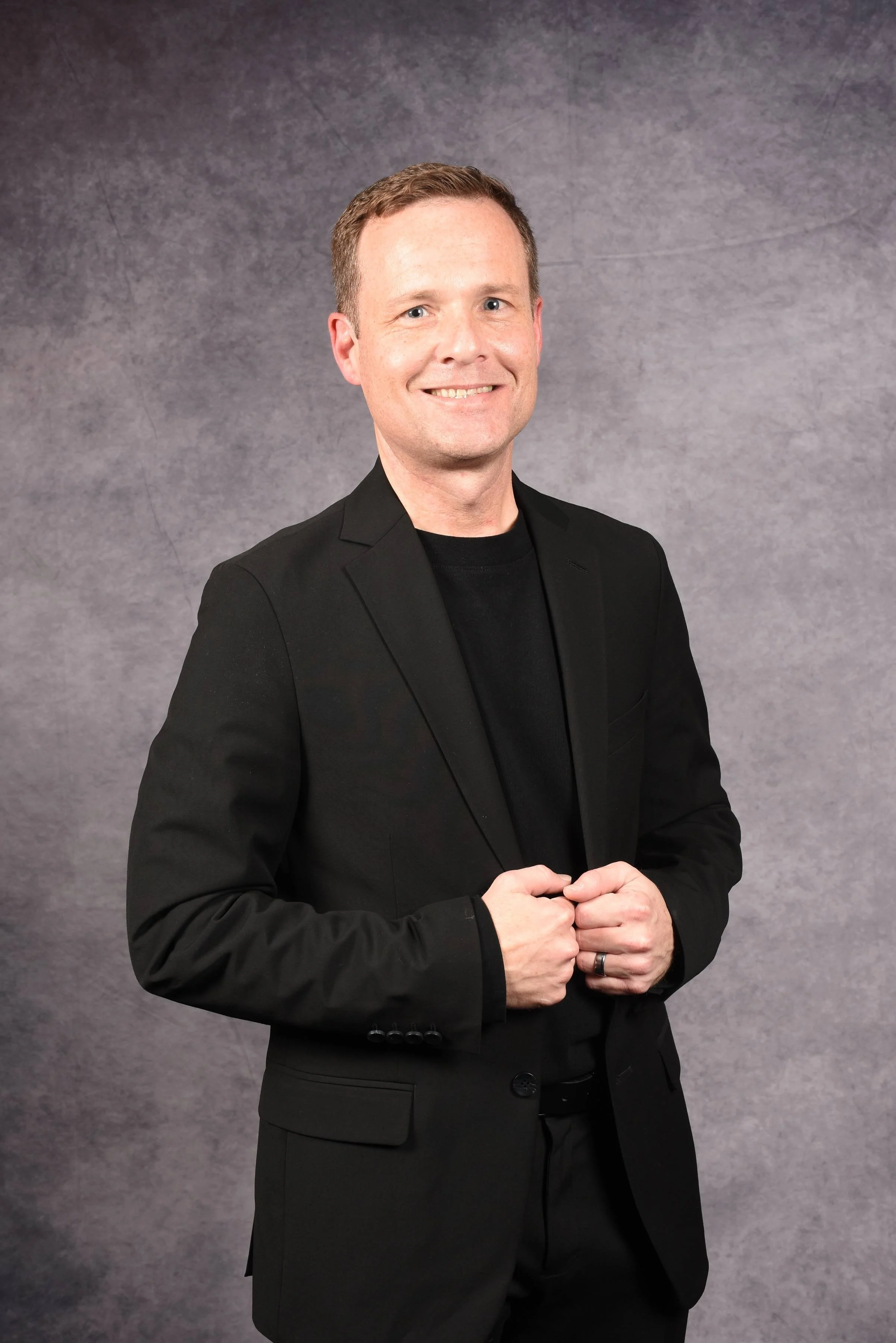 A man in a black suit and black shirt gesturing with his hands against a gray background.