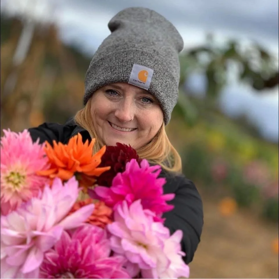 A woman wearing a lie gray Carhartt beanie smiling at the camera, holding a bunch of colorful dahlias in a garden or flower farm.