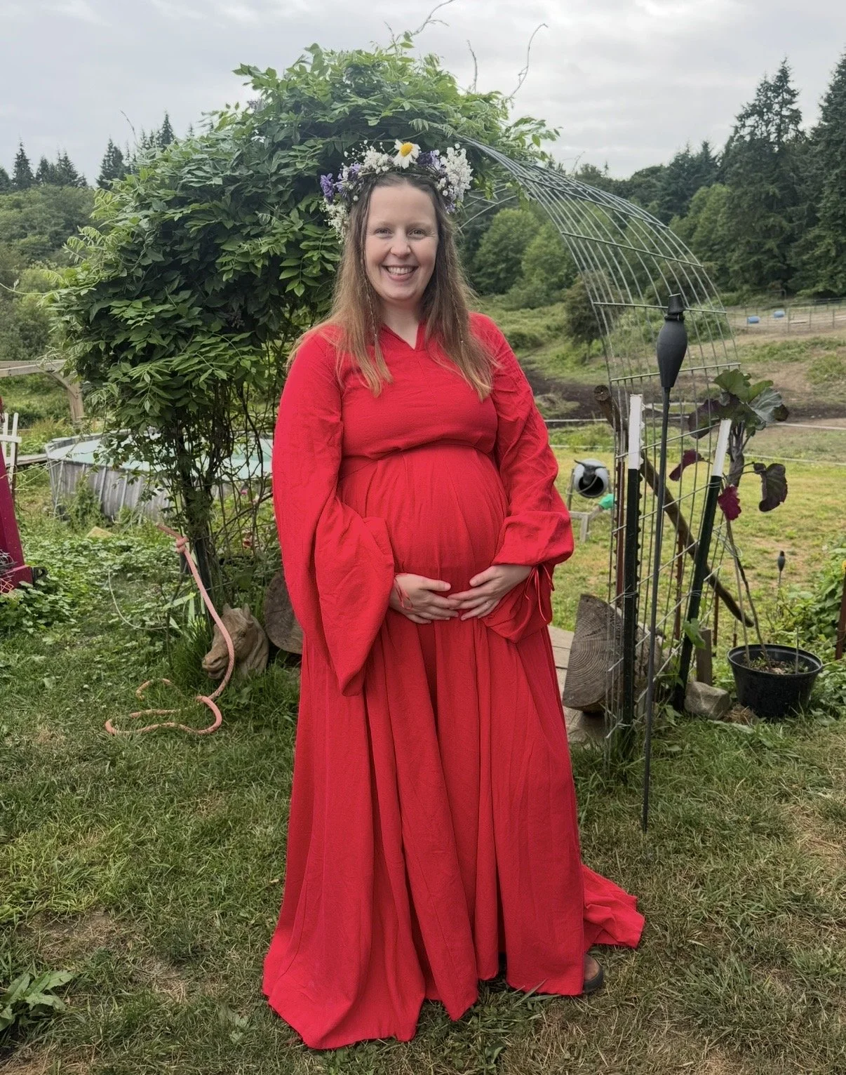 A pregnant woman in a long red dress with bell sleeves and a floral crown, standing outside in a garden.