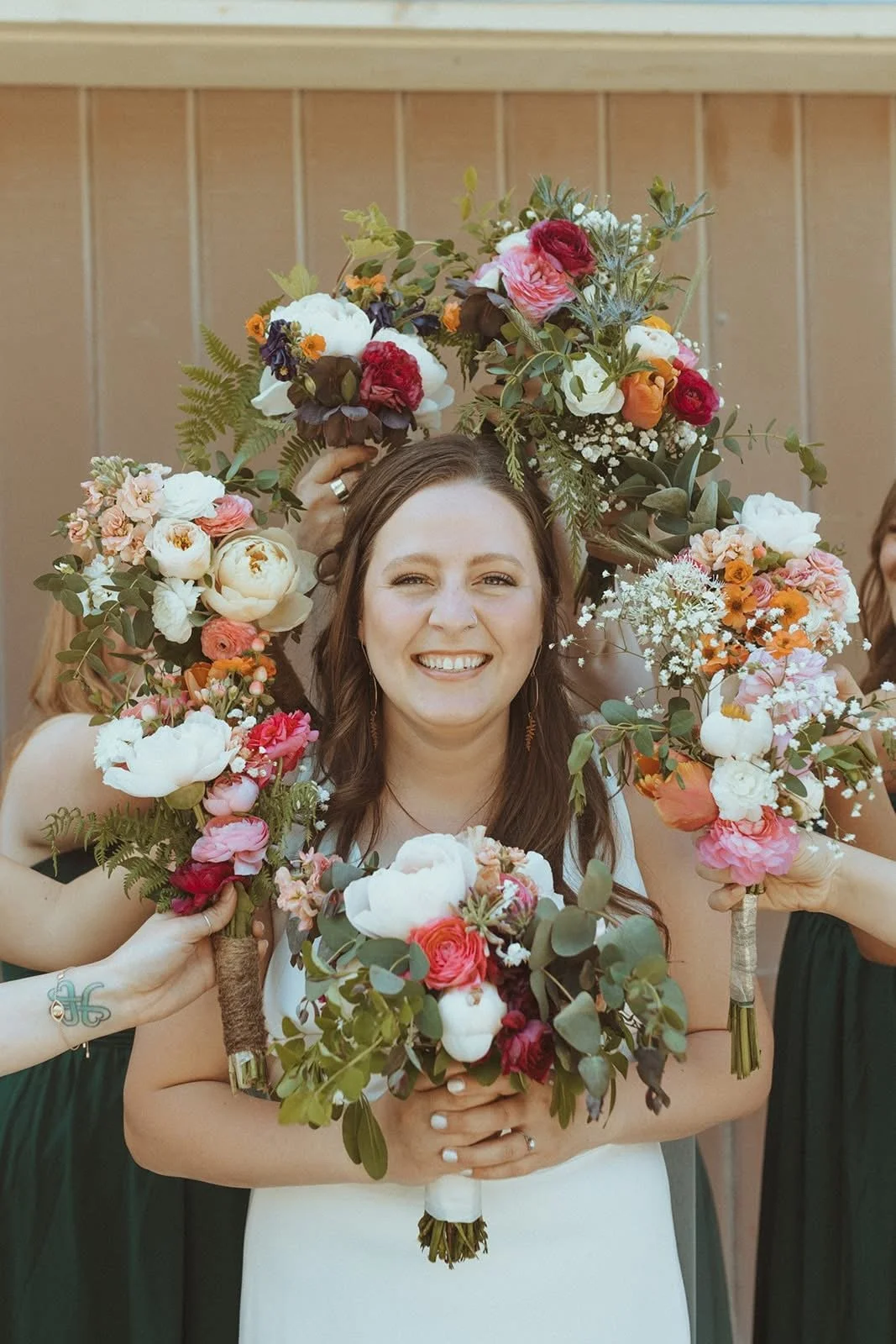 A woman smiling and holding a bouquet of colorful flowers surrounded by others holding floral arrangements. The background features wooden paneling.
