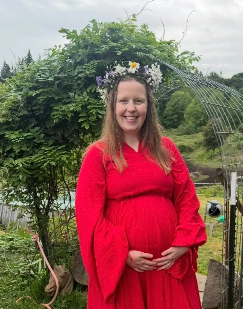 A pregnant woman wearing a red dress and a flower crown, standing outdoors in front of greenery and a garden arch, smiling and cradling her belly.
