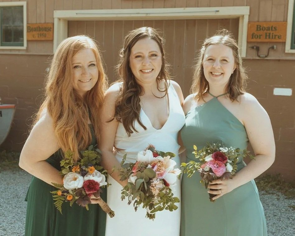 Three women standing outdoors, holding bouquets of flowers, smiling at the camera. The woman in the middle is wearing a white dress, and the other two are wearing green dresses. Behind them, a wooden building has signs that say "Barnhouse c. 1940" and "Boats for Hire."