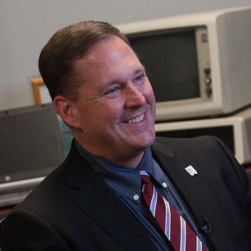 A smiling man in a dark suit and striped tie sitting in an office with computers in the background.