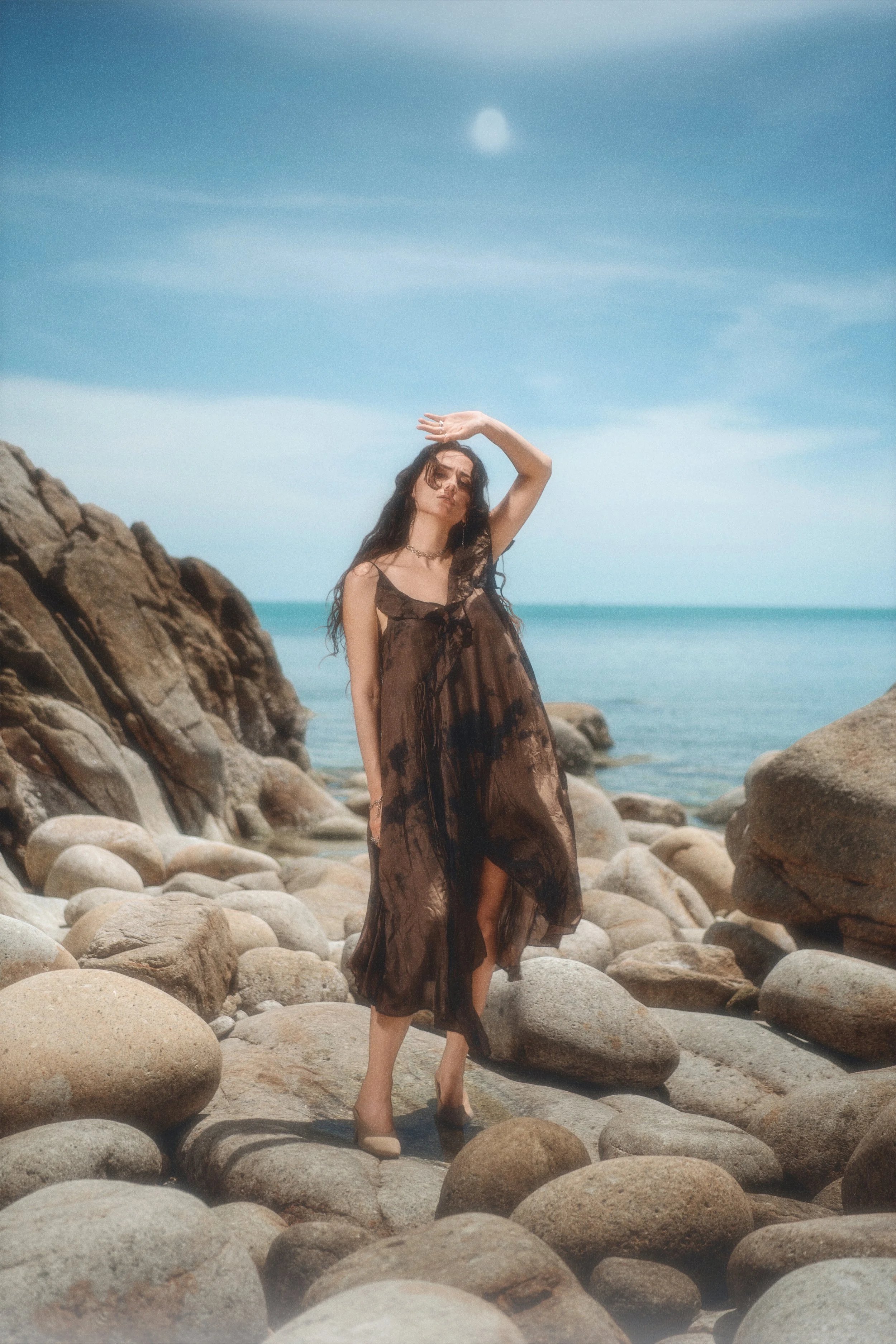 A woman in a see-through brown dress standing on a rocky beach with her hand raised above her head, rocks in the foreground, and a cloudy sky with the sun in the background.