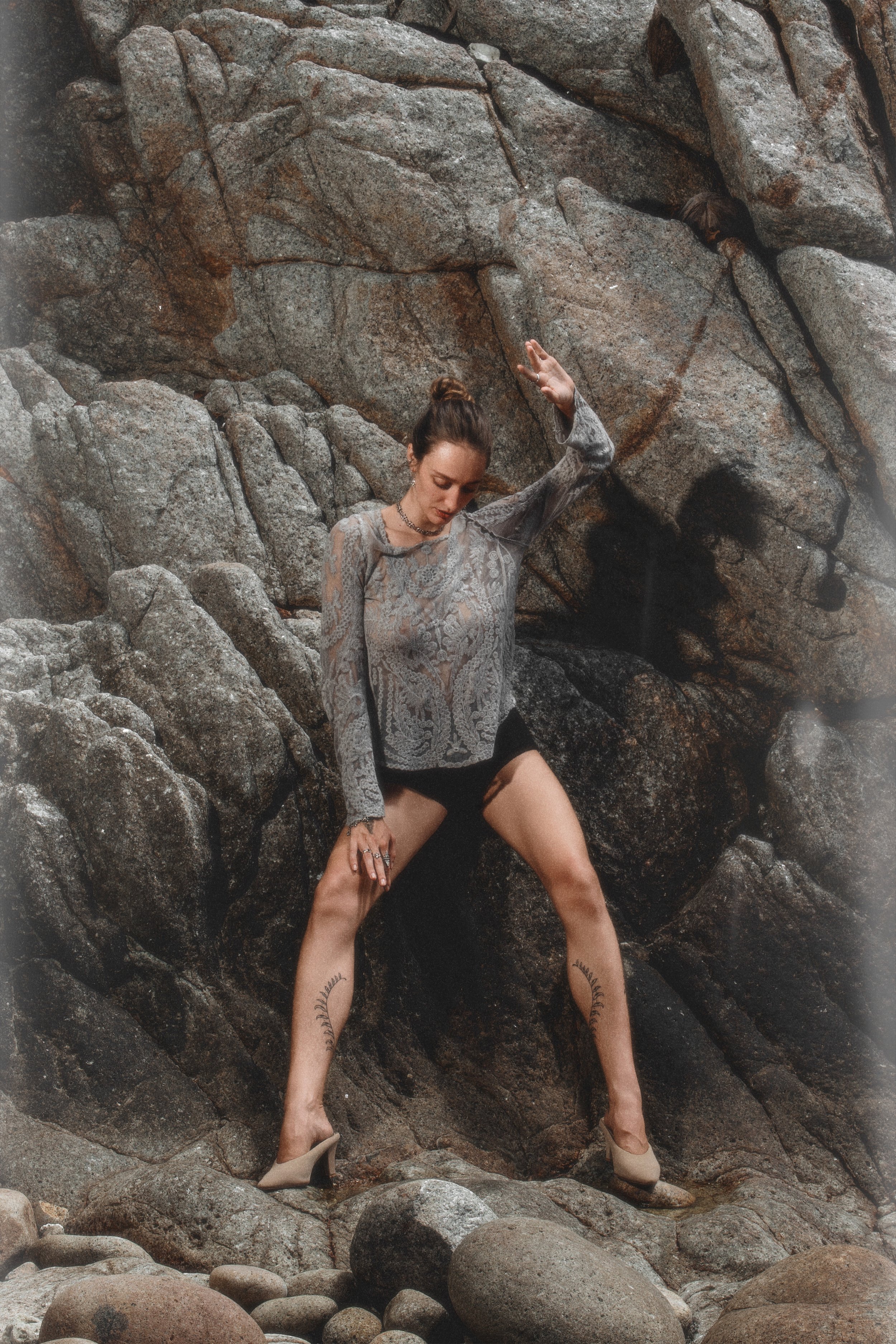 A woman with a top bun hairstyle is posing against a rocky background. She has tattoos on her legs and is wearing a lace top, black shorts, and beige high heels.