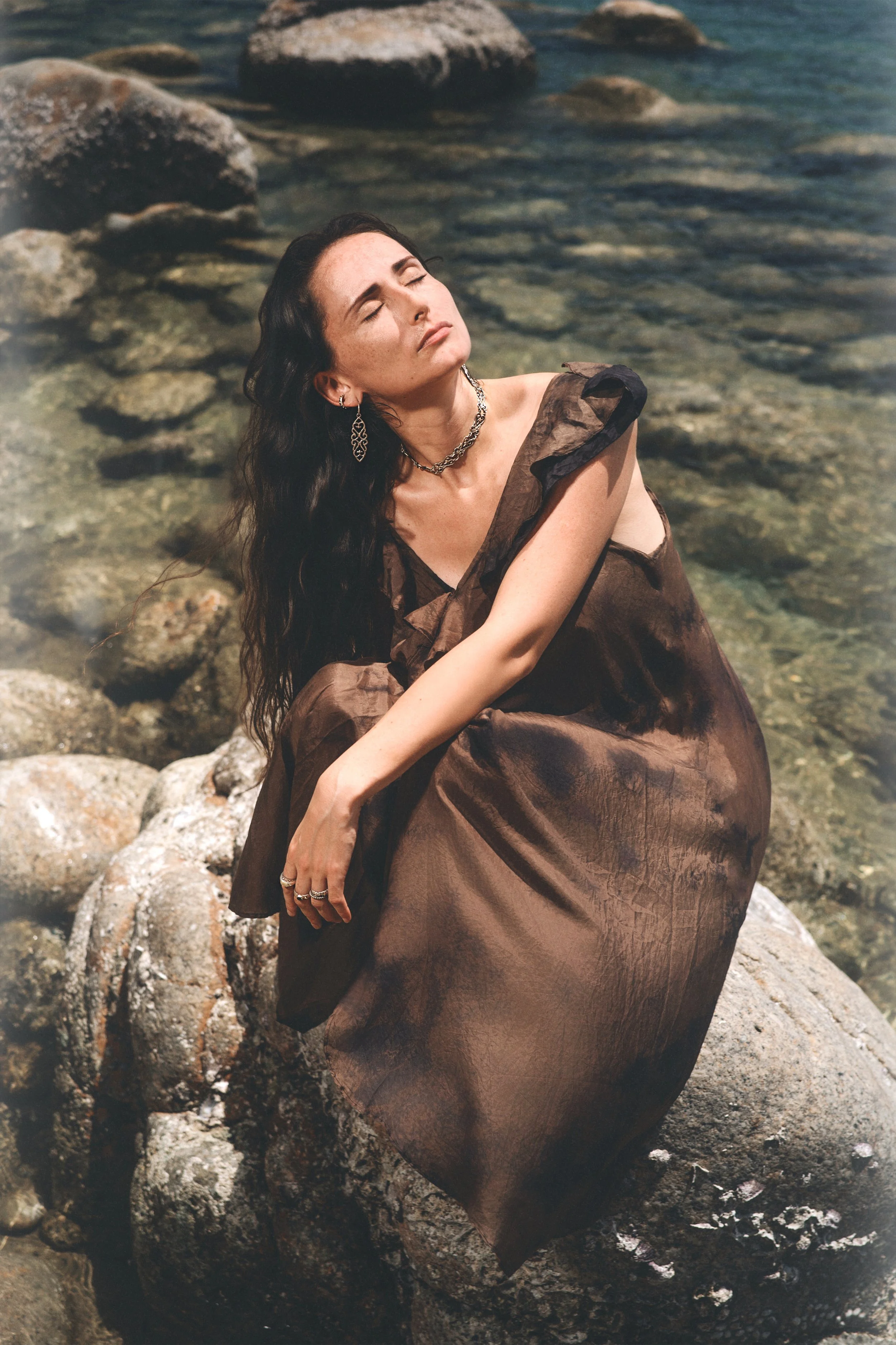 Woman with long dark hair, wearing silver jewelry and a brown dress, sitting on rocks by the water with eyes closed.