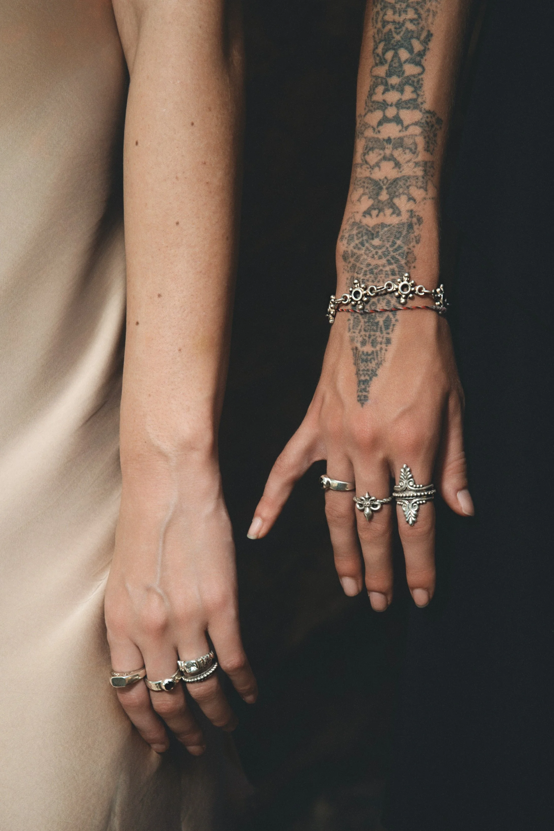 Two hands with multiple rings and bracelets resting against each other, one is tattooed and wearing a silver bracelet, the other is uninked.