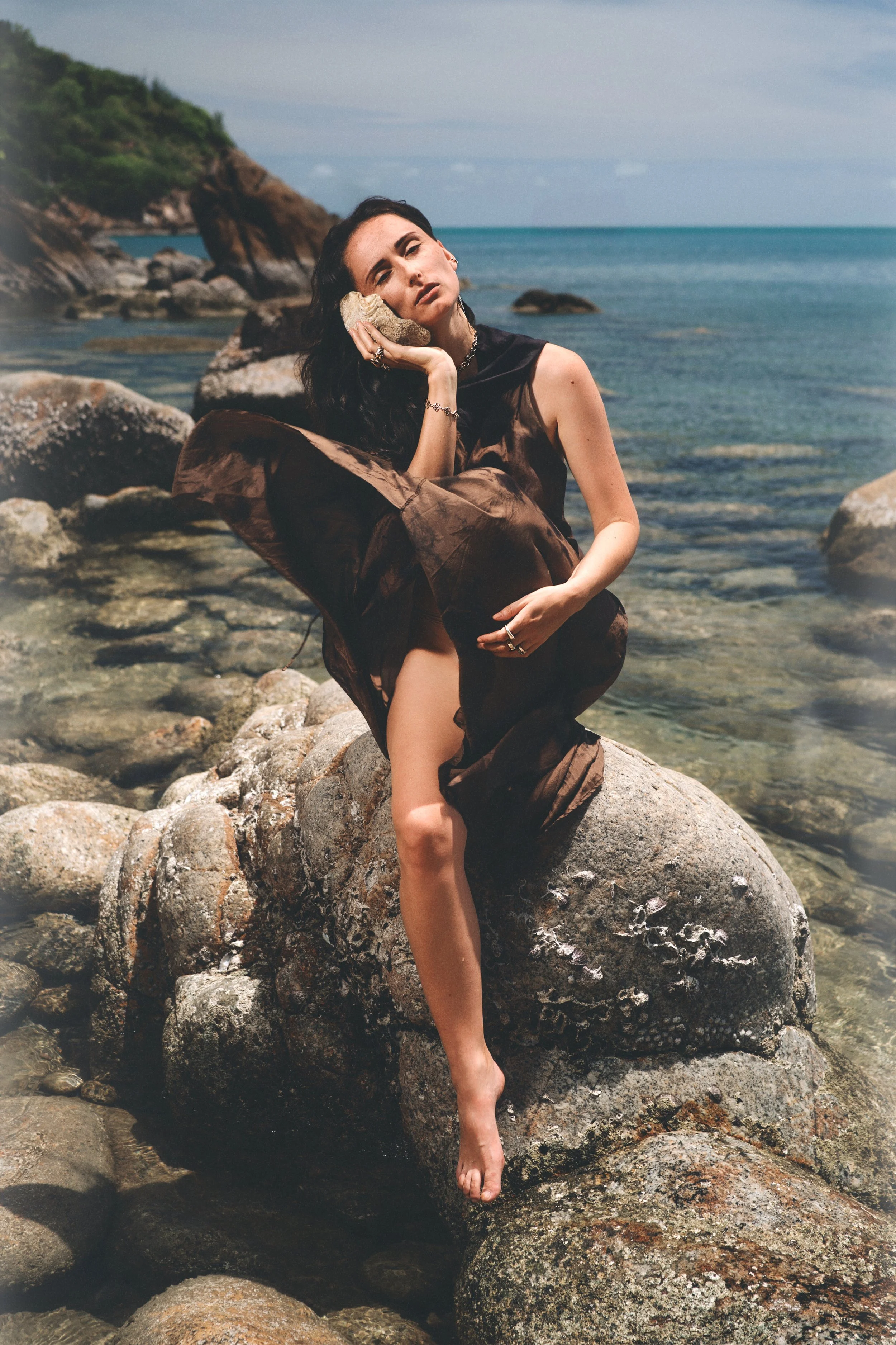 A woman with dark hair sits on a large rock on a beach, resting her head on a rock in her hand, with her body partially covered by a semi-transparent brown fabric. The ocean and a rocky coastline are visible in the background.