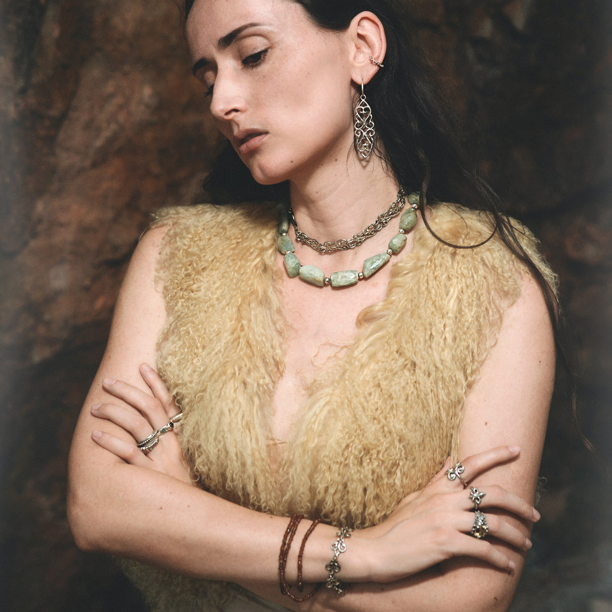 A woman with dark hair is wearing multiple jewelry pieces including earrings, necklaces, rings, and bracelets, and is dressed in a fuzzy beige vest with a brown background.