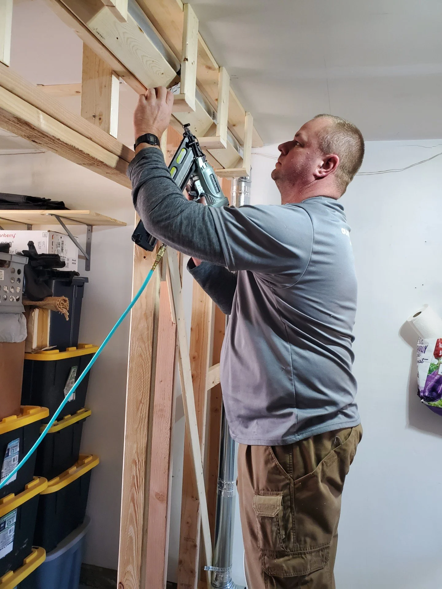 A man in a gray long sleeve shirt and tan cargo pants is installing or working on a wooden framework, using a pneumatic nail gun in a garage or workshop setting.