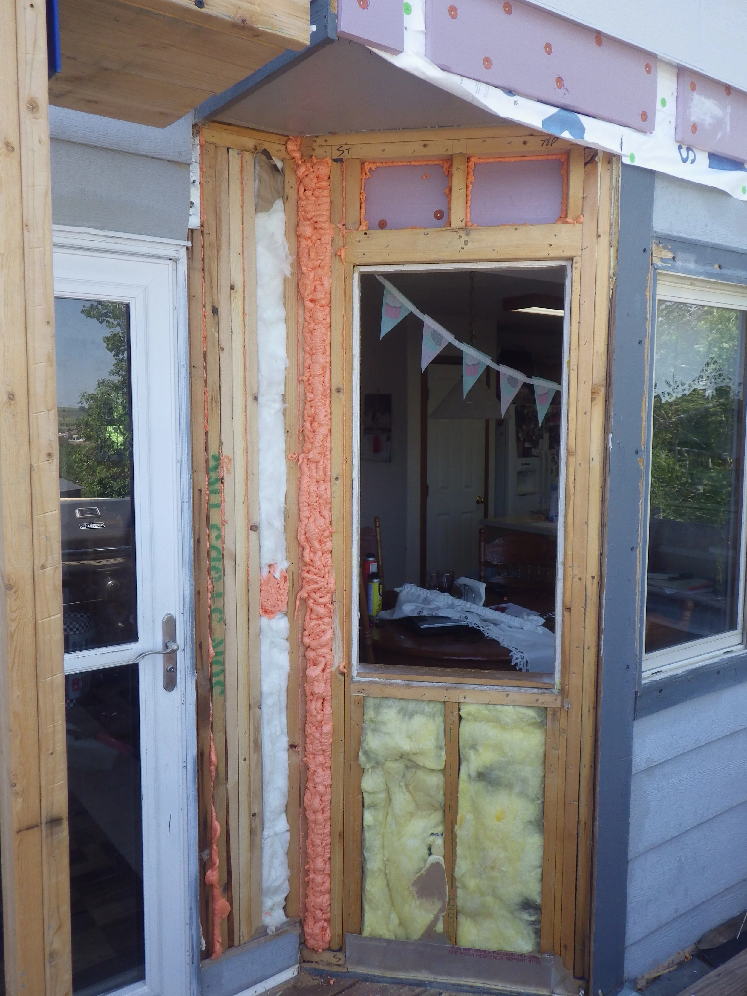External view of house undergoing renovation with exposed wooden framing, insulation, and partial installation of a storm door and window.