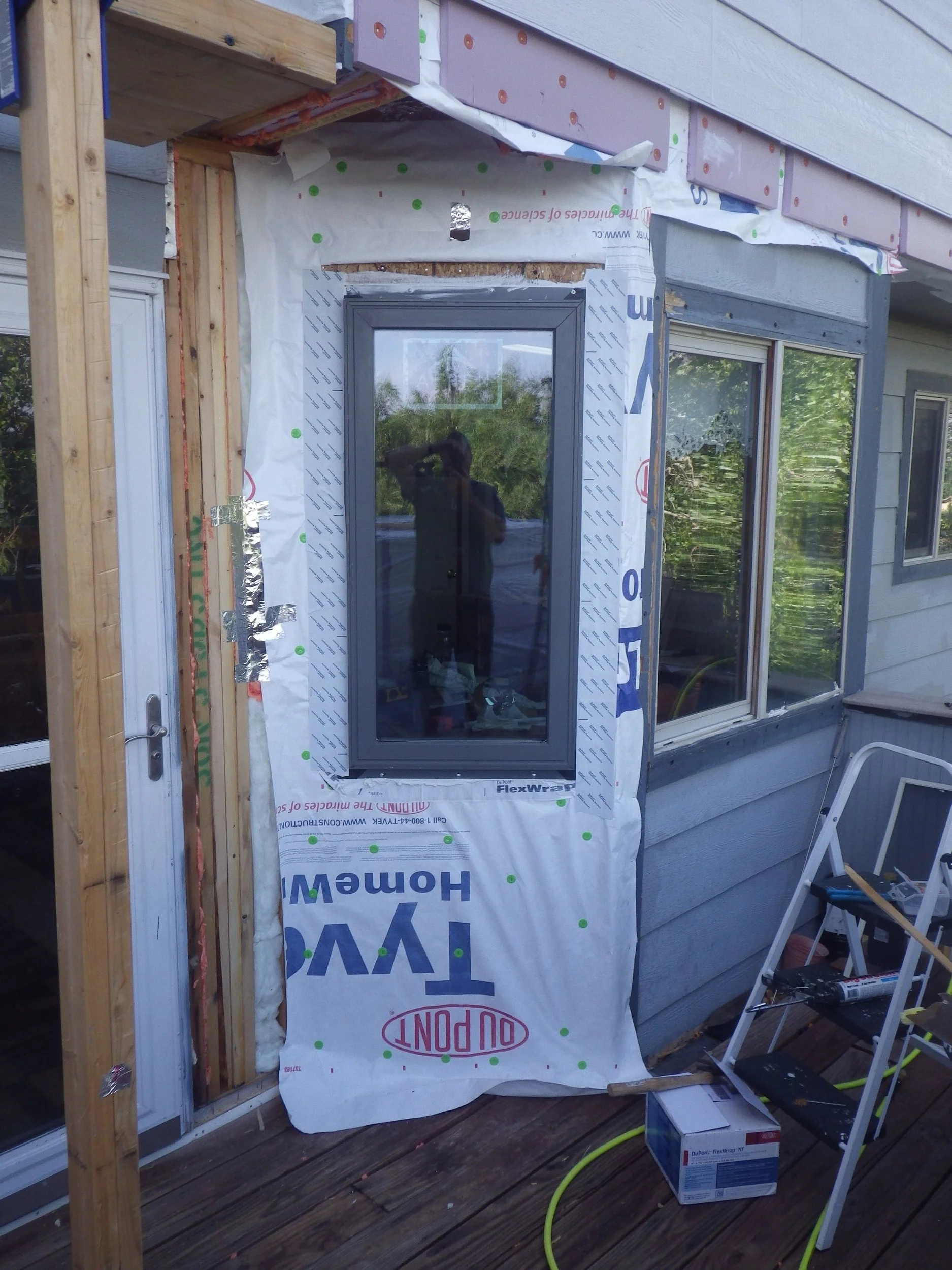 Construction site showing partial exterior of a house with a new window installation, building materials, and tools on a wooden deck.