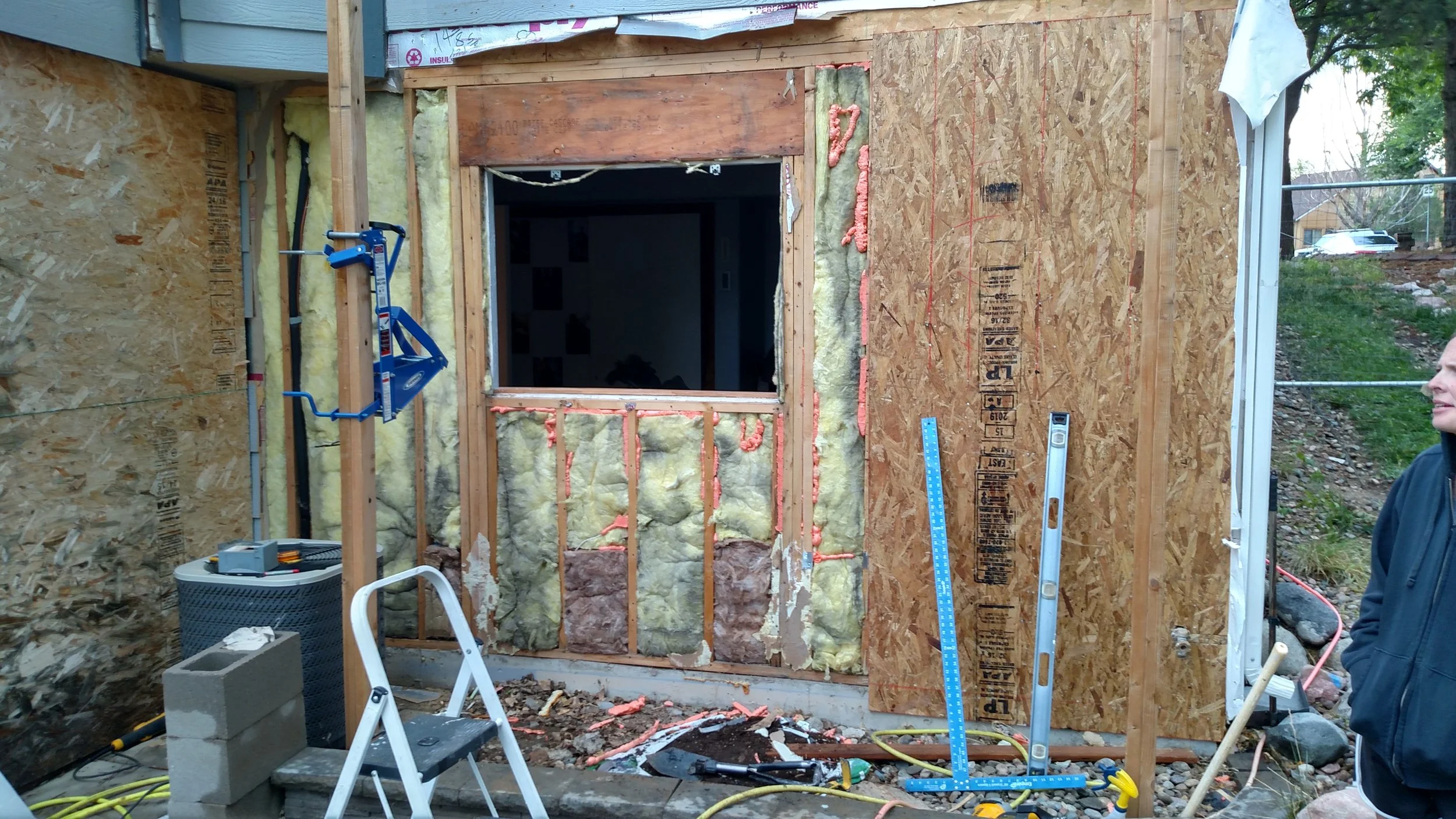 Construction site with unfinished wall, insulation, window opening, ladder, and tools; person standing nearby