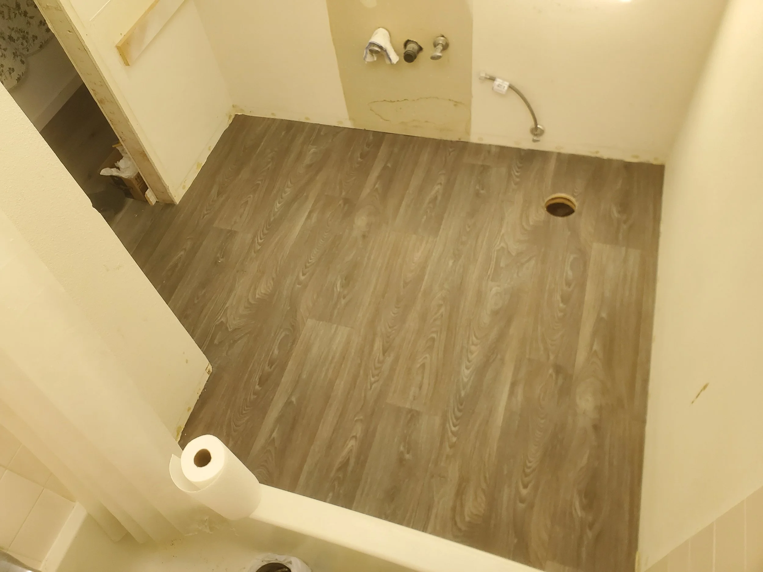 Empty bathroom with new vinyl flooring, plumbing pipes without fixtures, and a floor drain hole.
