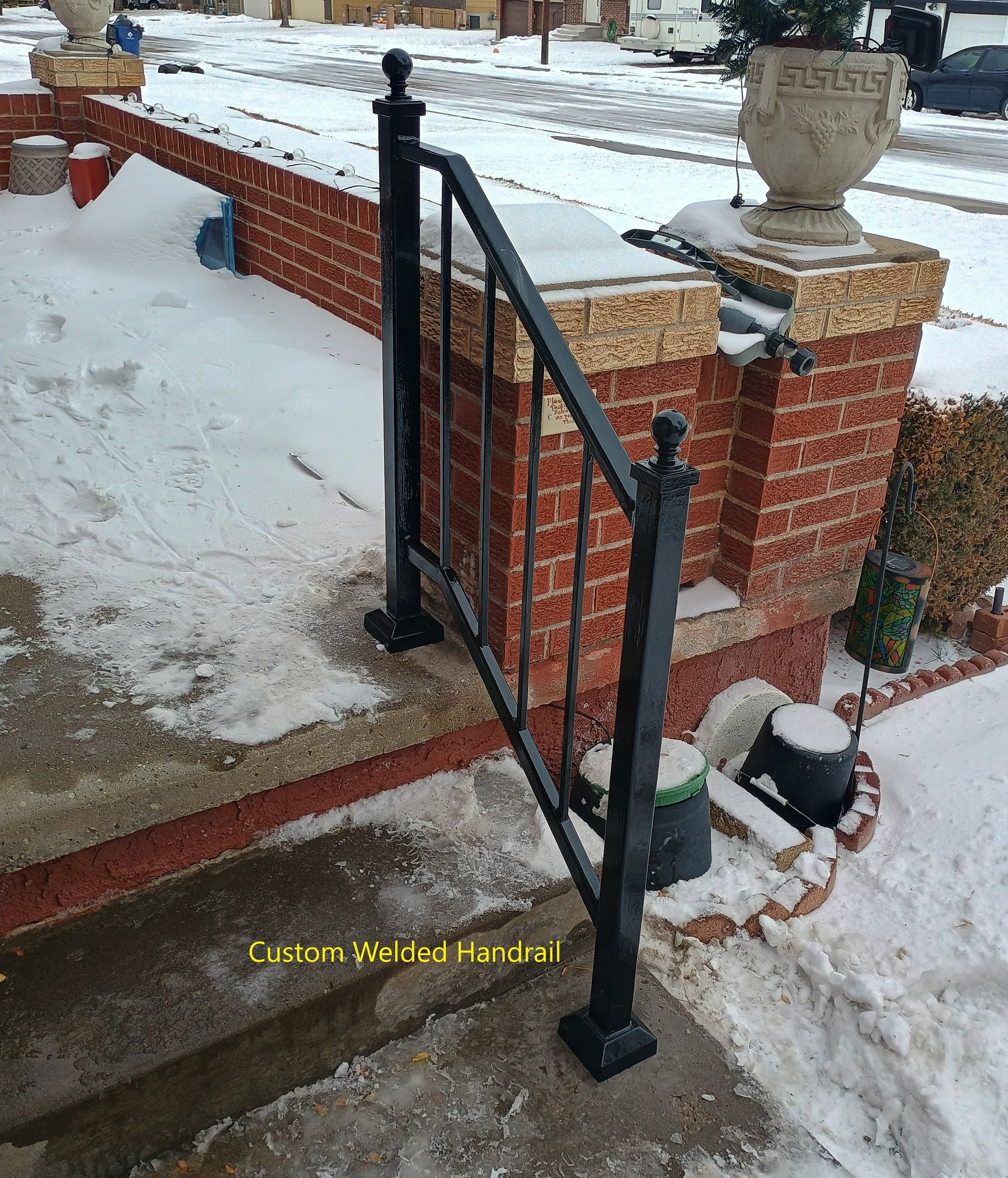 Close-up of a black, custom welded handrail installed on concrete steps outside a brick house, with snow on the ground and decorative pots and garden items nearby.