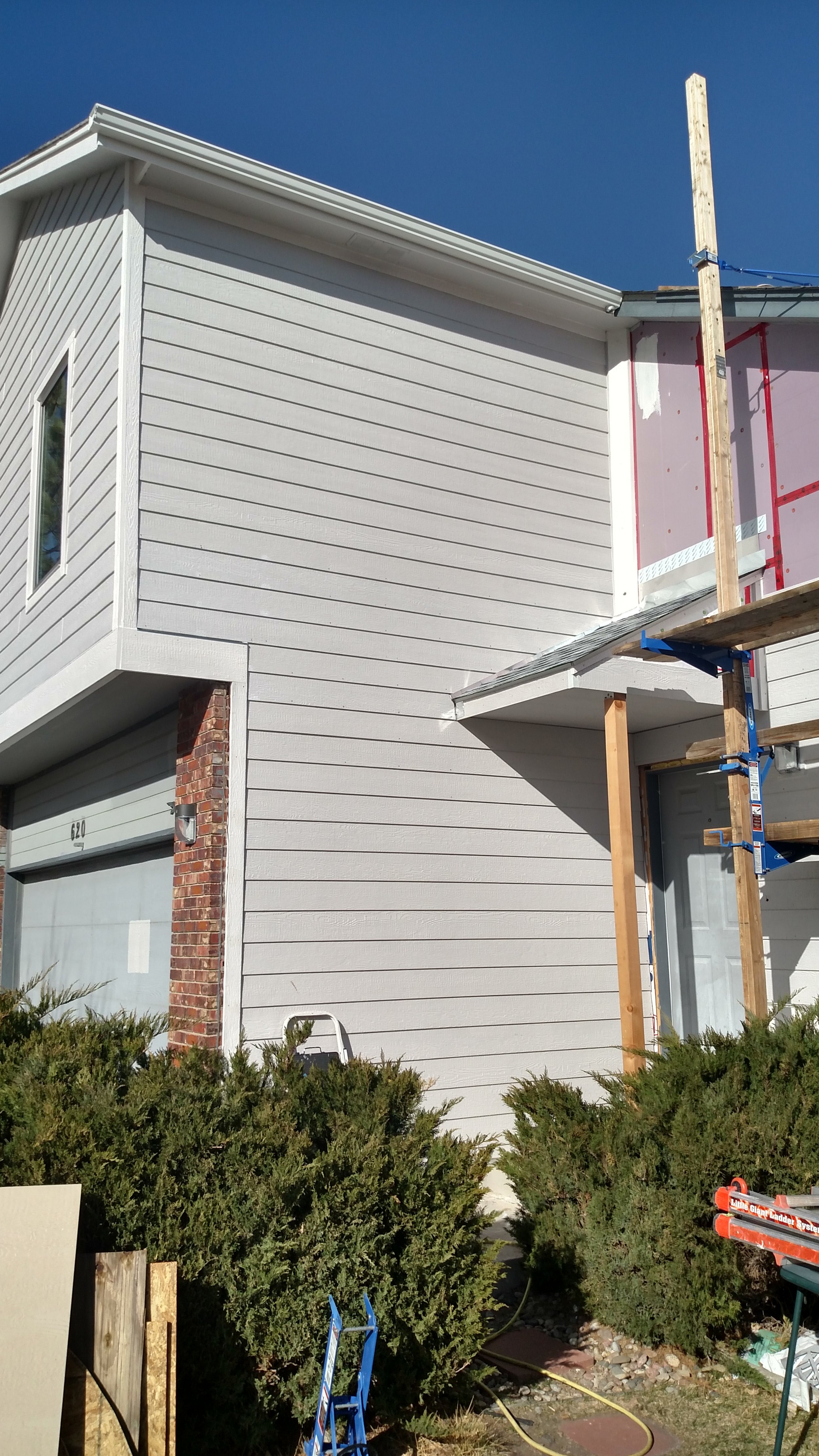 Front view of a house under construction with new white siding, a brick column, and scaffolding on the right side, with bushes in the foreground and a clear blue sky overhead.