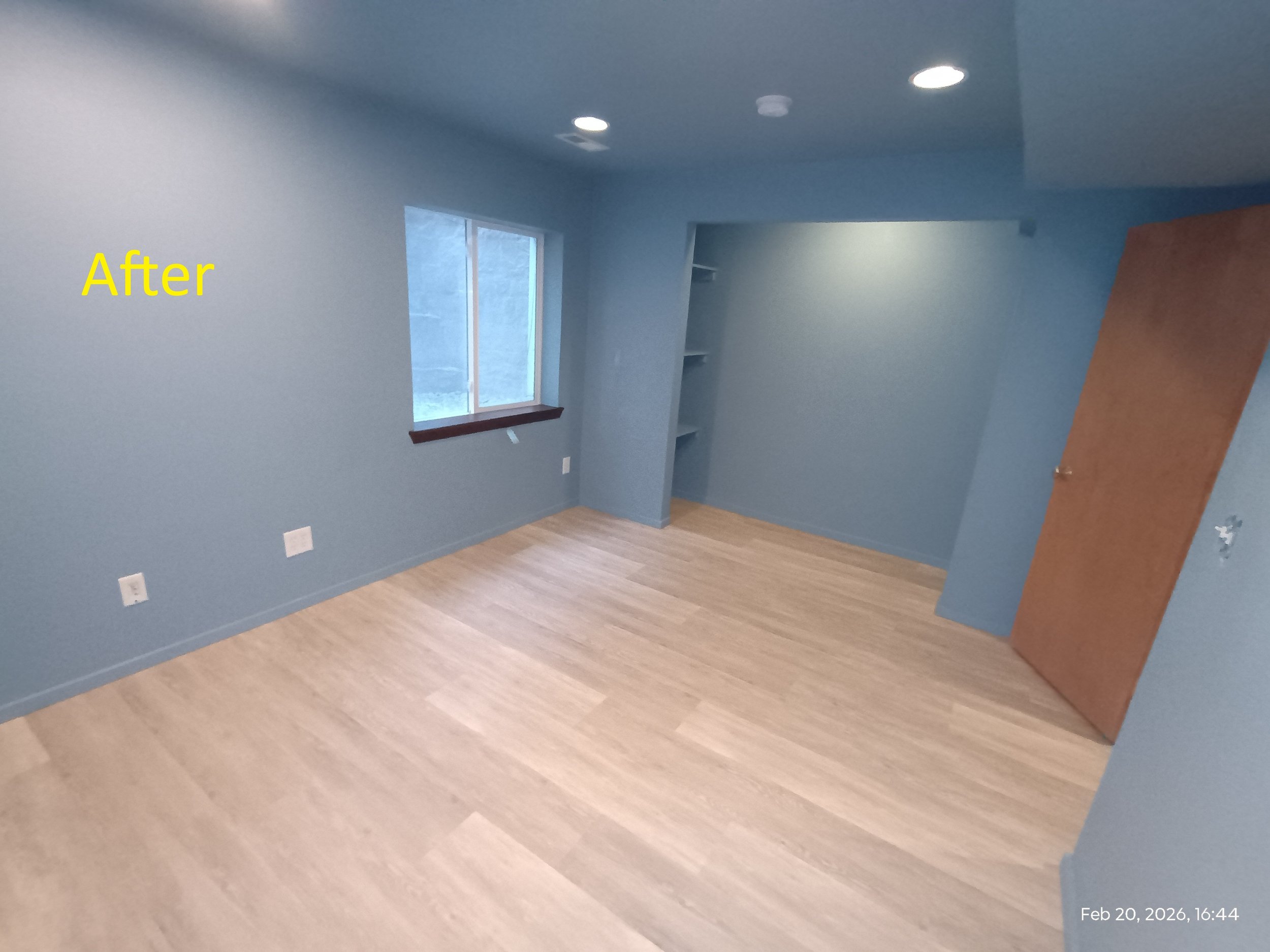 Empty room with blue walls, wooden flooring, small window, built-in shelves, and a closed wooden door with timestamp Feb 20, 2026, 16:44.