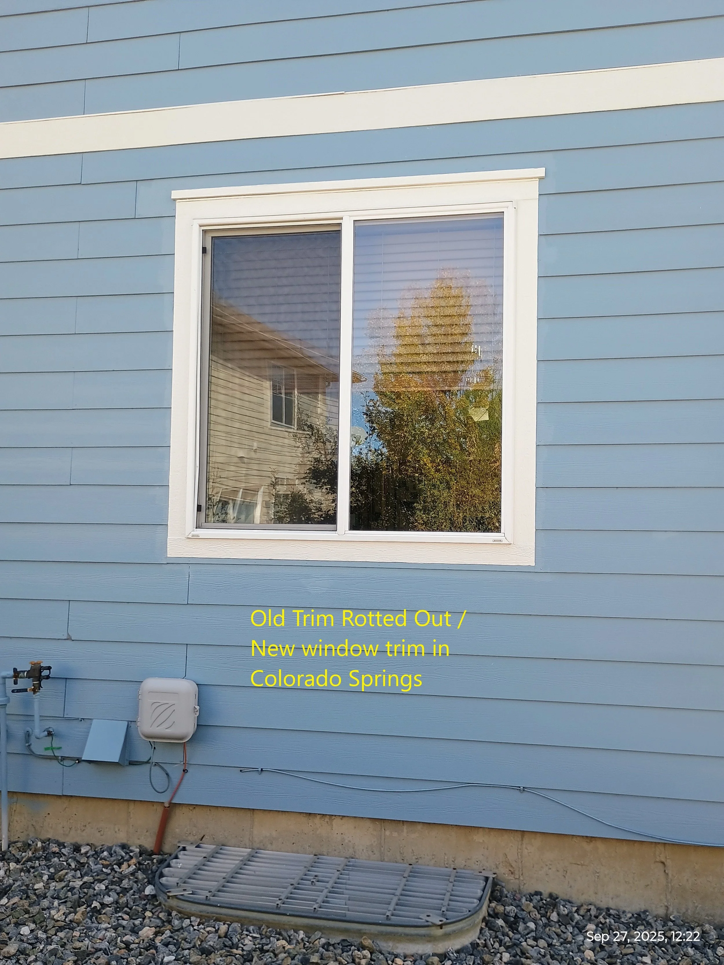 Close-up of a blue house exterior with a single white-trimmed window reflecting trees and neighboring buildings. The wall has horizontal siding, and there are utility connections and a window air conditioning unit below. Yellow text notes the old win