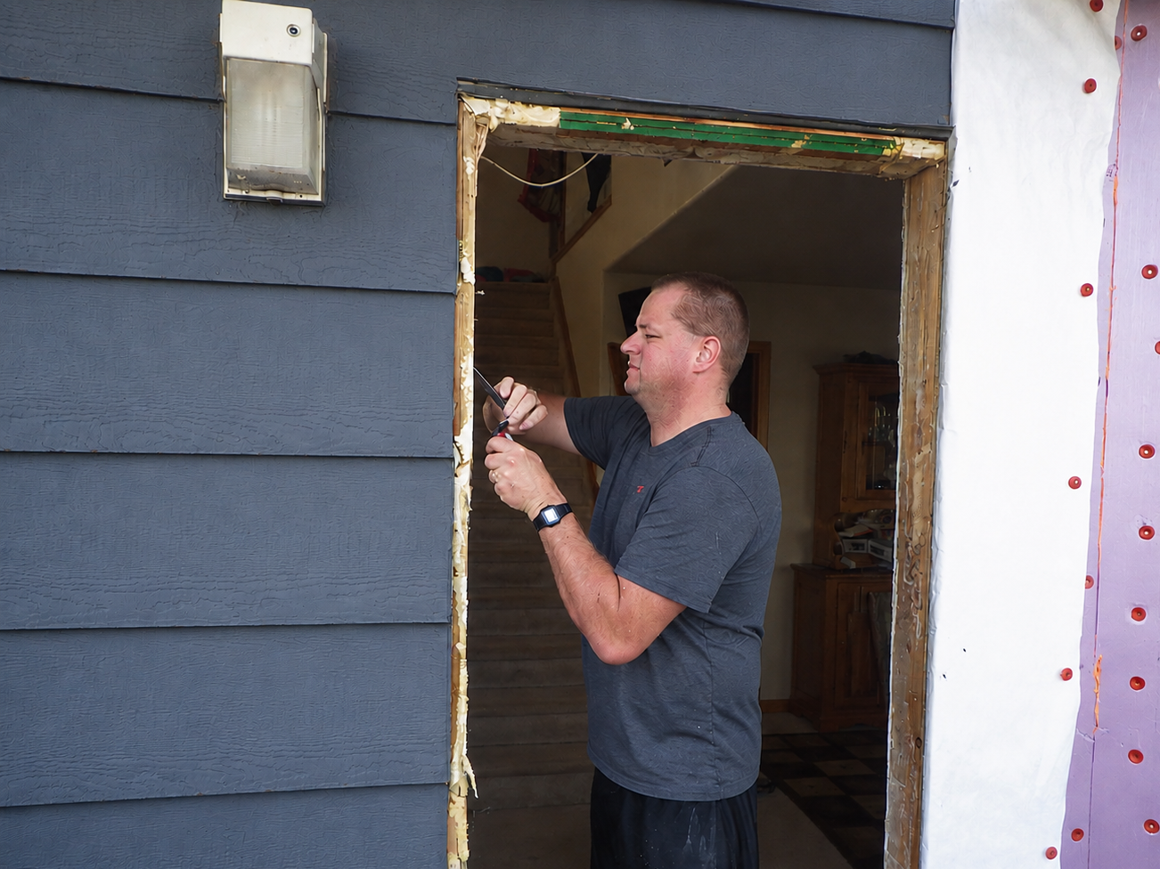A man with a smartwatch is using a screwdriver on the frame of a house under renovation, with house siding and insulation visible.