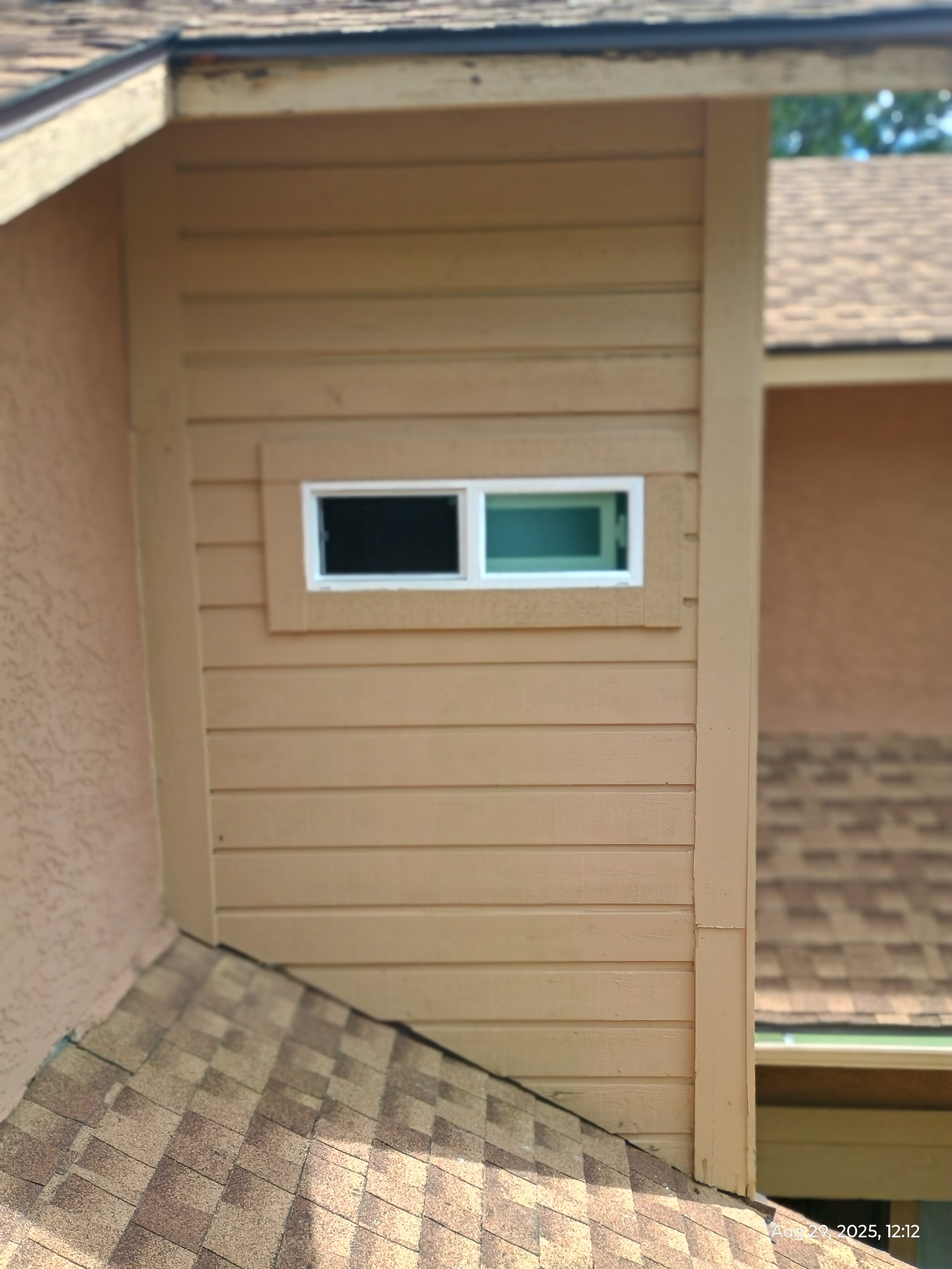Photo of an exterior corner of a building with beige horizontal wooden siding, a white window, and a brick archway on the right side. The ground is paved with multicolored bricks, and part of a roof with shingles is visible at the top. The photo is timestamped August 29, 2025, 12:12.
