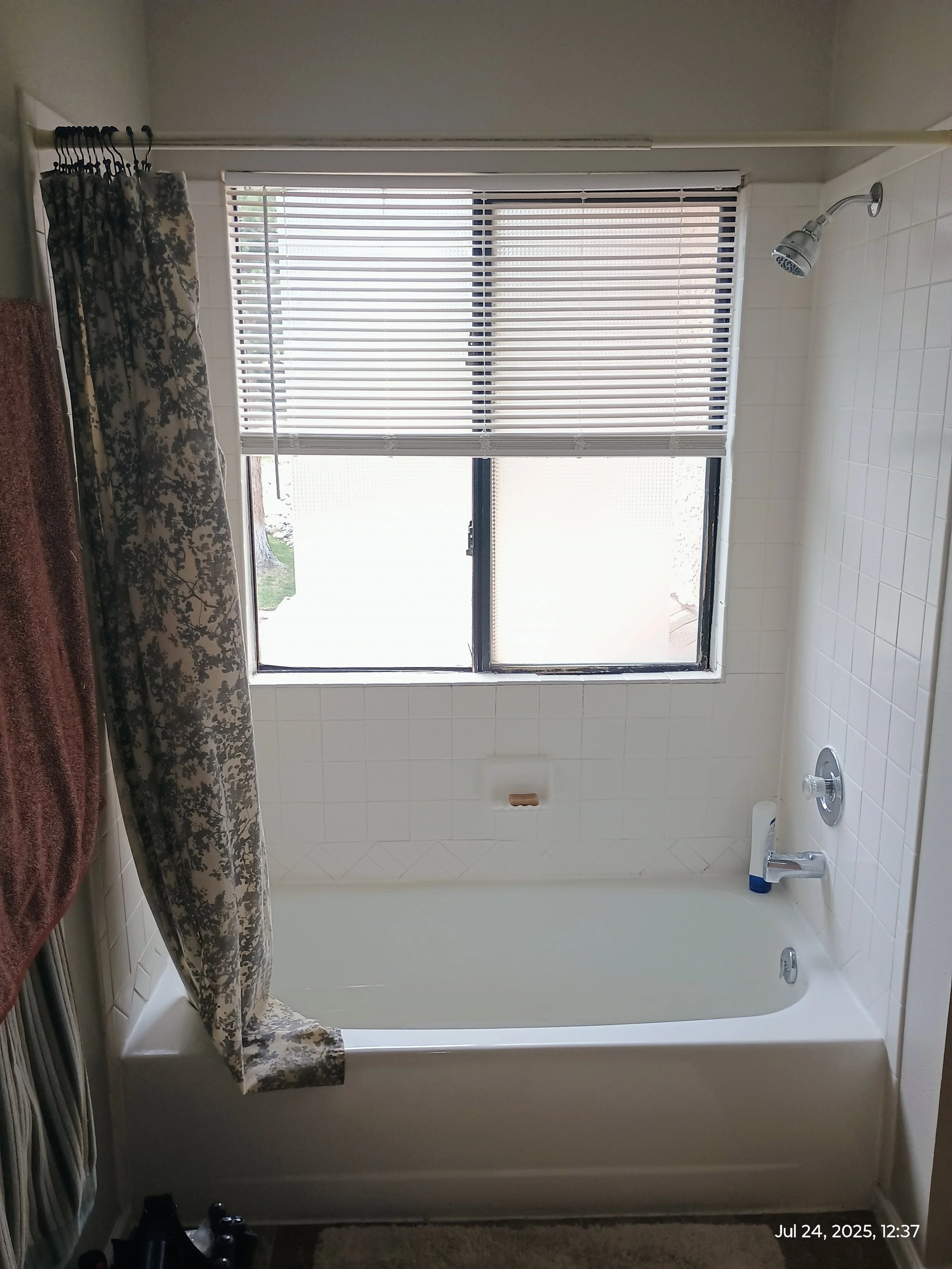 A bathroom with a bathtub and shower, a window with blinds, a patterned curtain, and some toiletries near the bathtub.