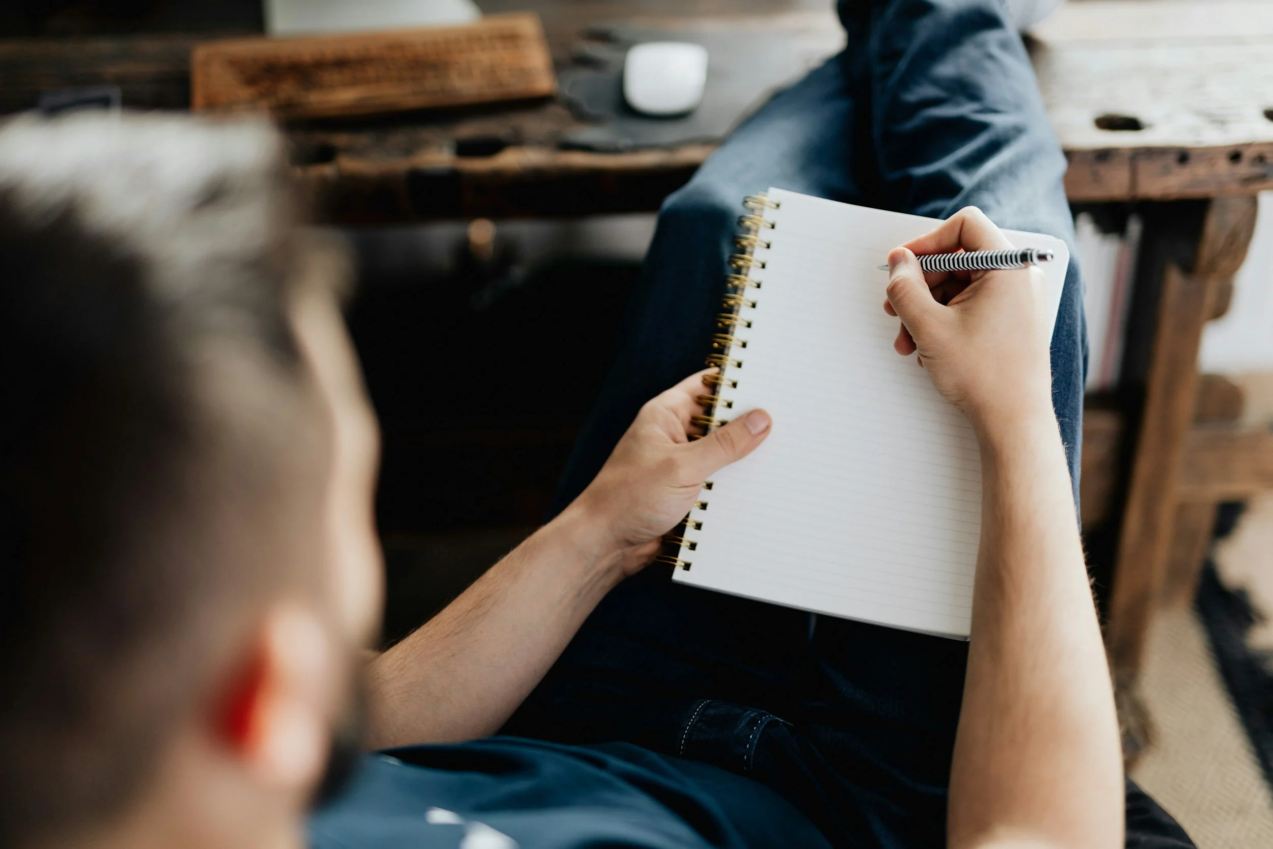 Person sitting cross-legged on the floor taking notes in a spiral notebook with a black and white pen, with a wooden table and a white speaker in the background.
