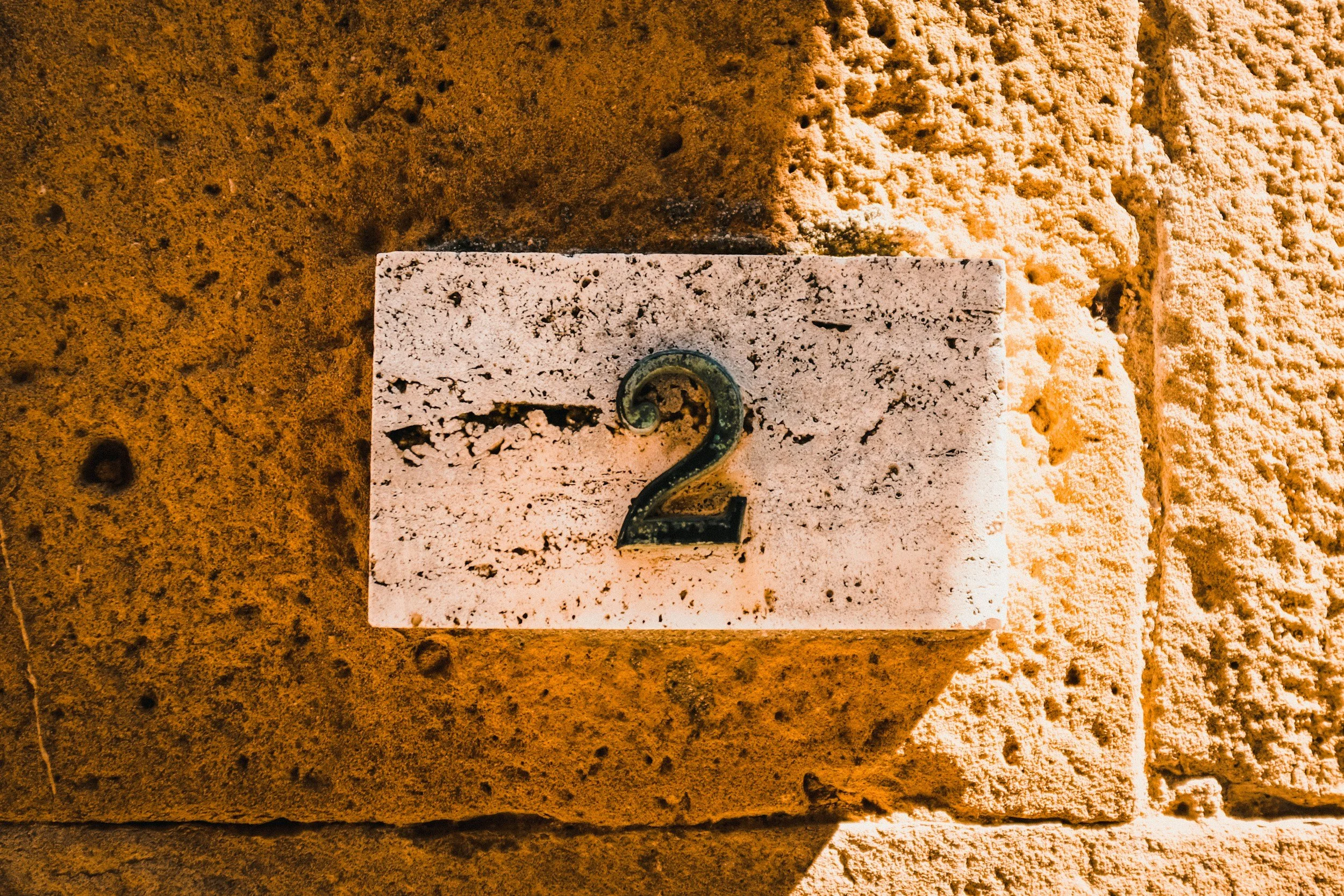 A close-up of a textured brick wall with a small white square plaque featuring a black number 2 and a green question mark.