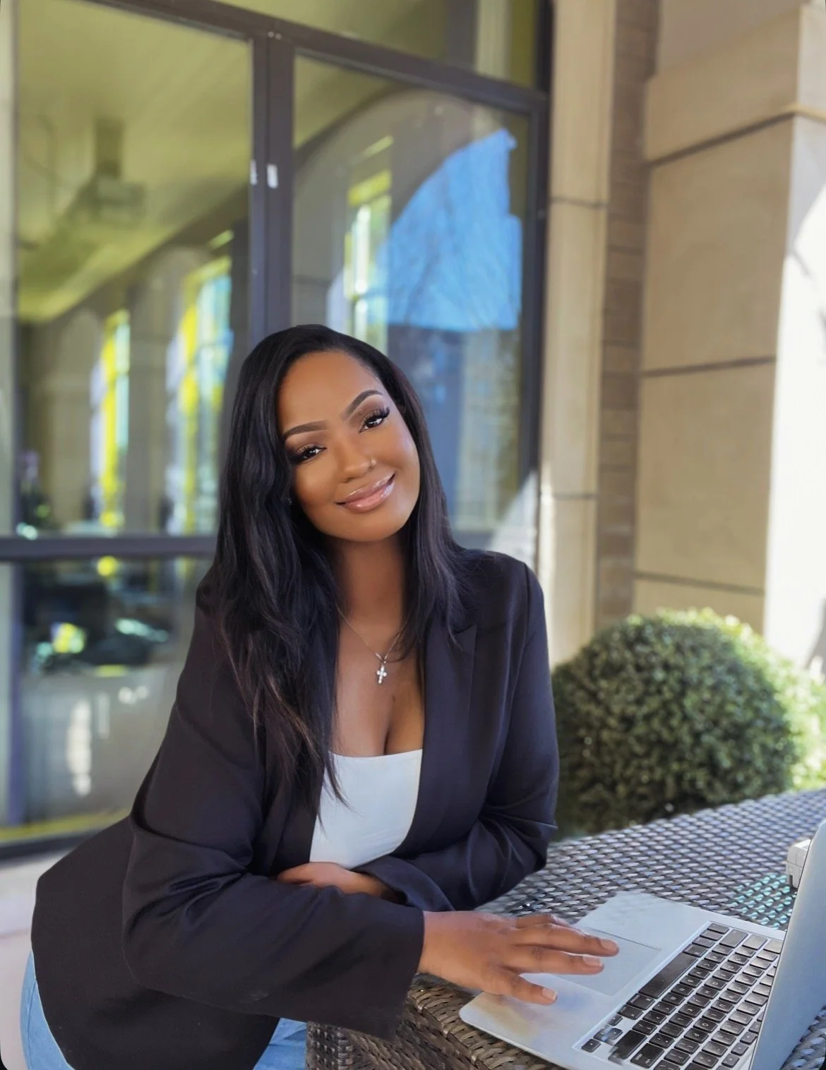 A woman sitting outdoors at a table with a laptop, smiling and wearing a black blazer and white top, with a building and window in the background.