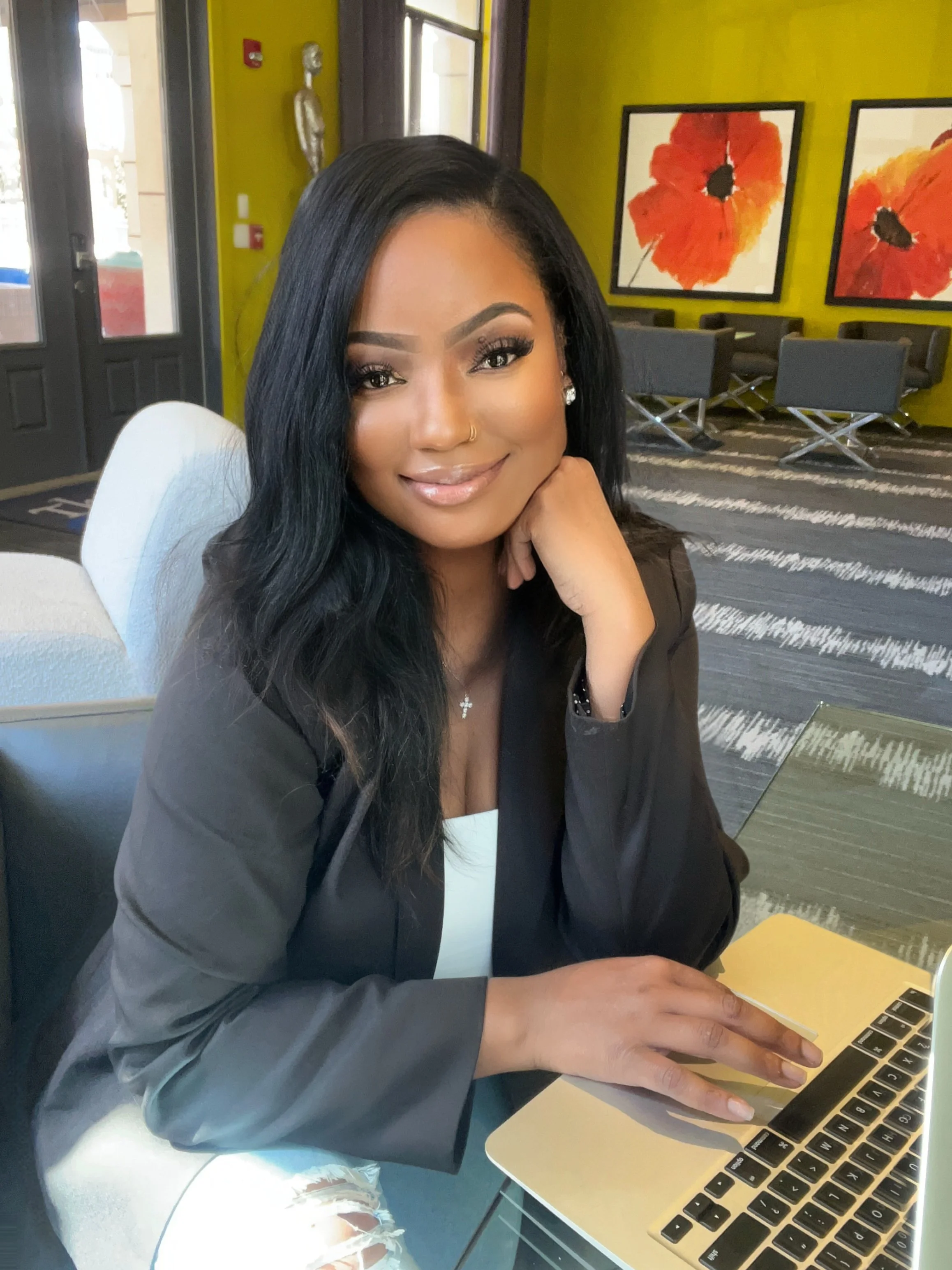 A woman with long black hair, makeup, and jewelry, sitting at a table using a laptop in a colorful cafe or restaurant with green walls, floral artwork, and gray chairs.
