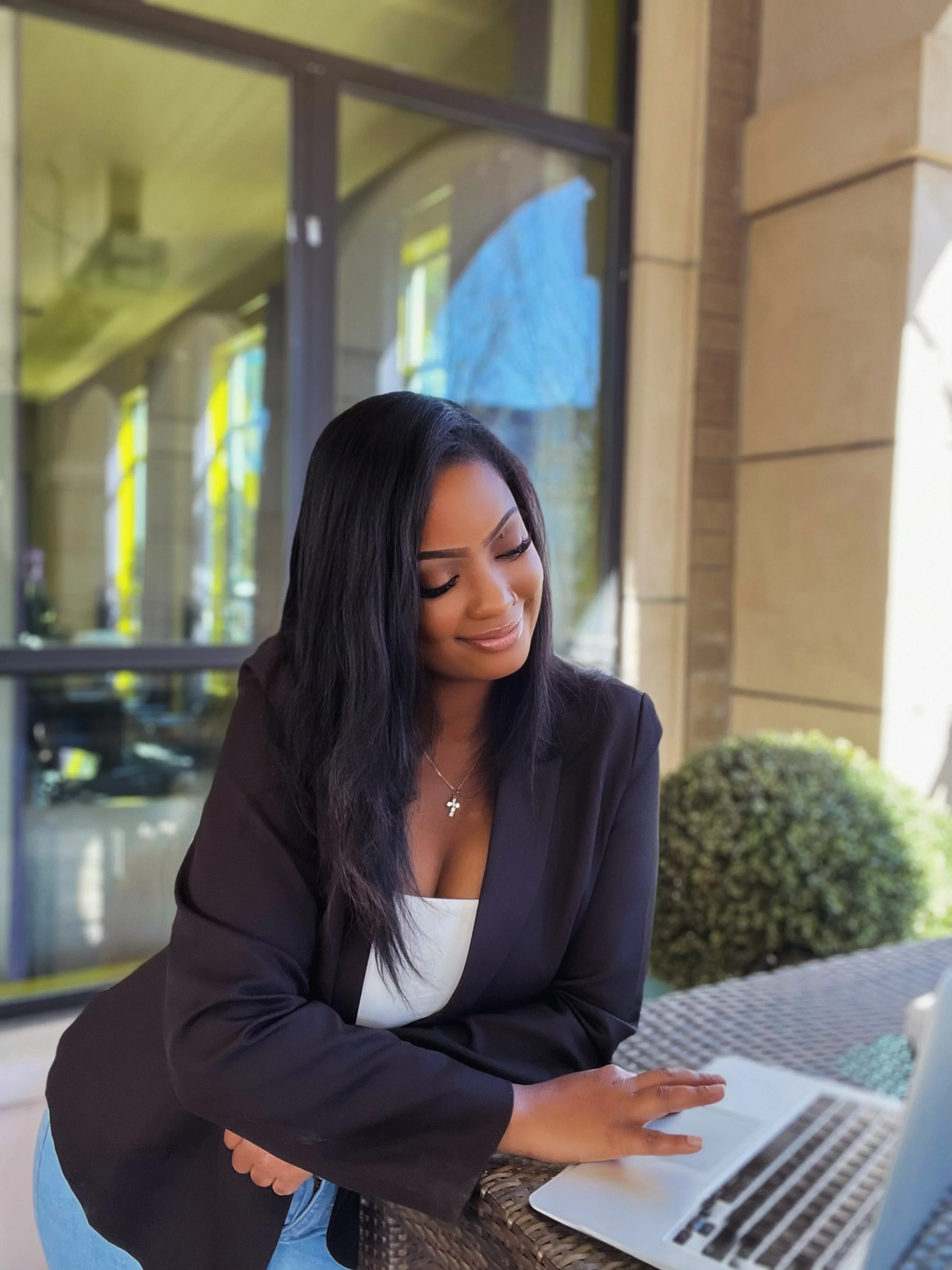 A woman with long black hair and a cross necklace working on a laptop at an outdoor cafe table.
