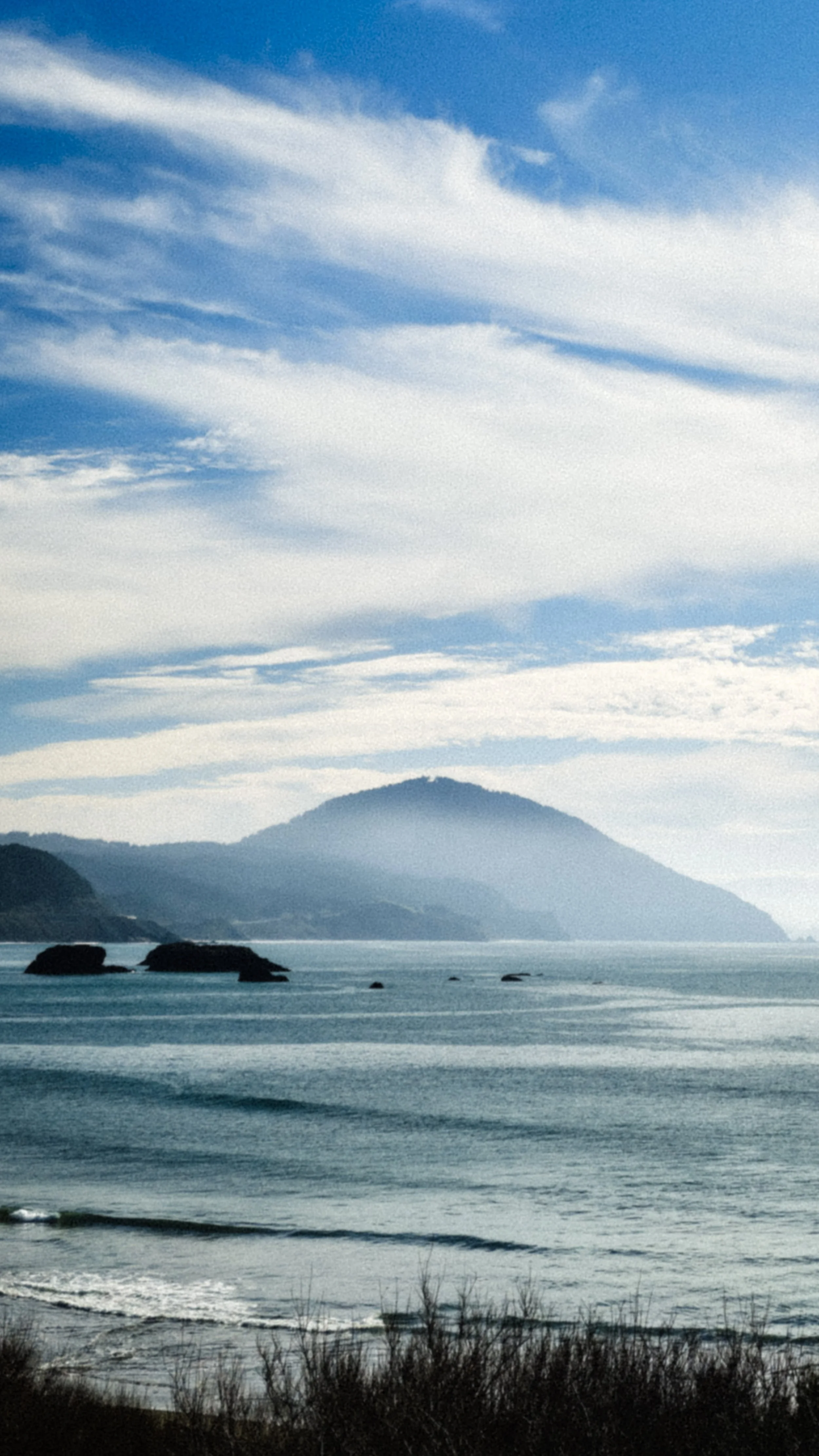 A scenic view of a large body of water with waves, rocky islands in the foreground, and a mountain in the background under a partly cloudy sky.