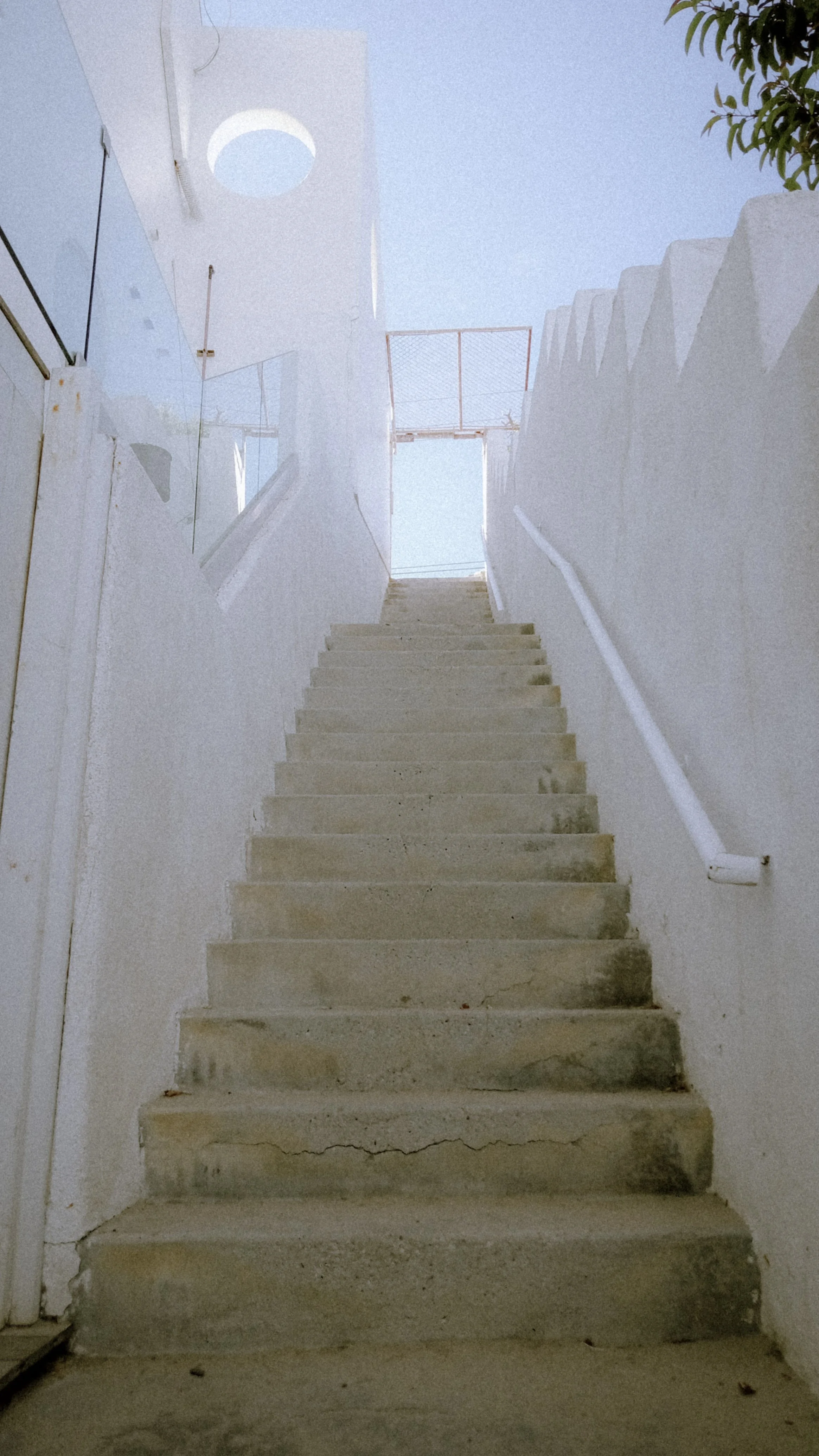 Concrete stairs leading upward with white walls on either side, a white handrail on the right, and a door or opening at the top with a metal gate. The sky is visible above.