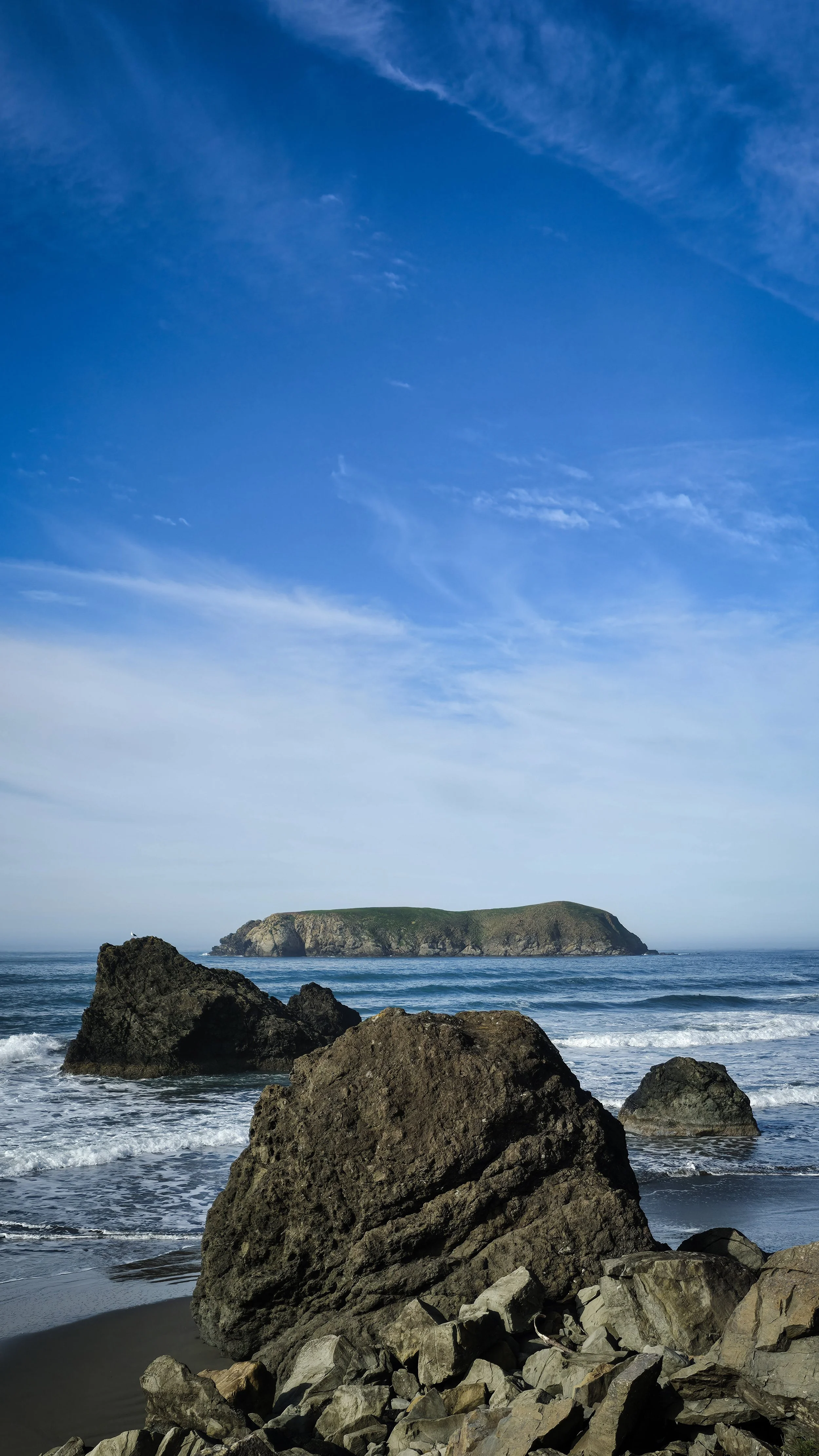 Rocky beach with large rocks in foreground, ocean waves, and a green island in the distance under a partly cloudy blue sky.