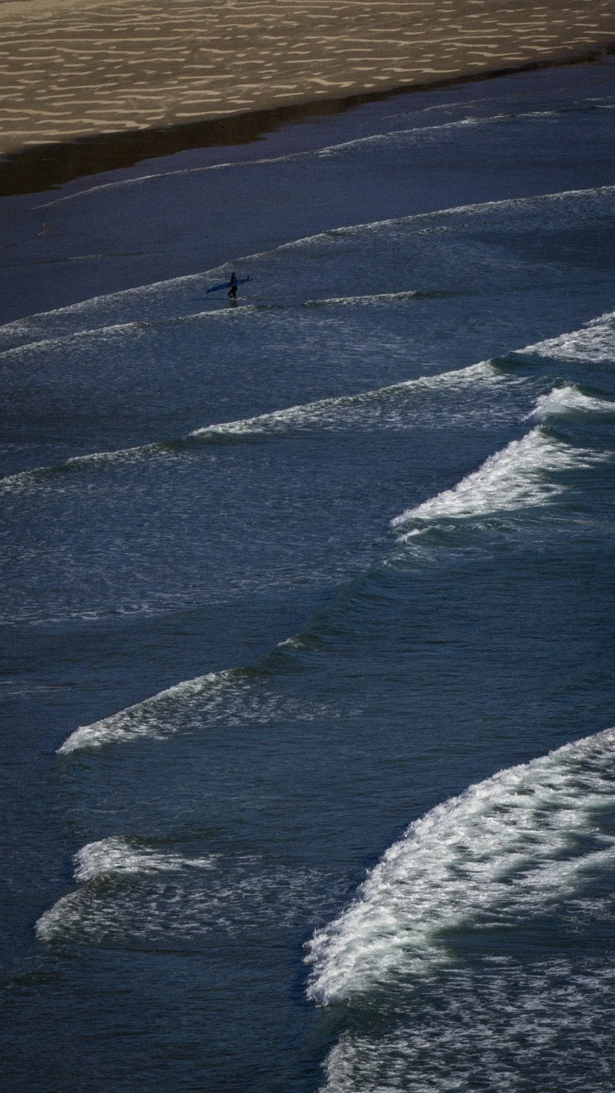 A person carrying a surfboard walking along the shoreline as waves break on the sandy beach at sunset.