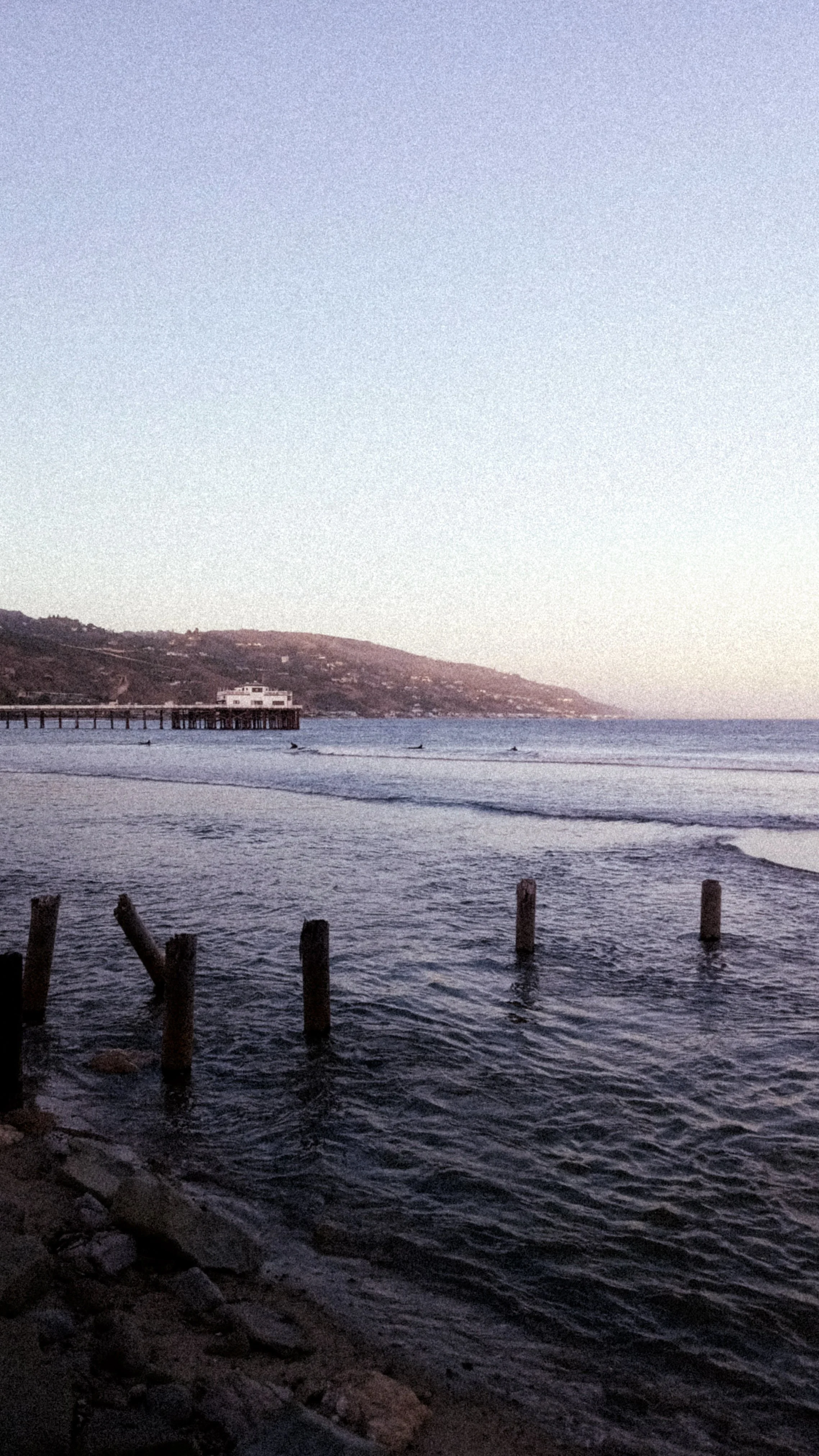 Sunset over the ocean with a rocky shoreline in the foreground, wooden posts in the water, and a pier with a white building extending into the water, hills in the background.