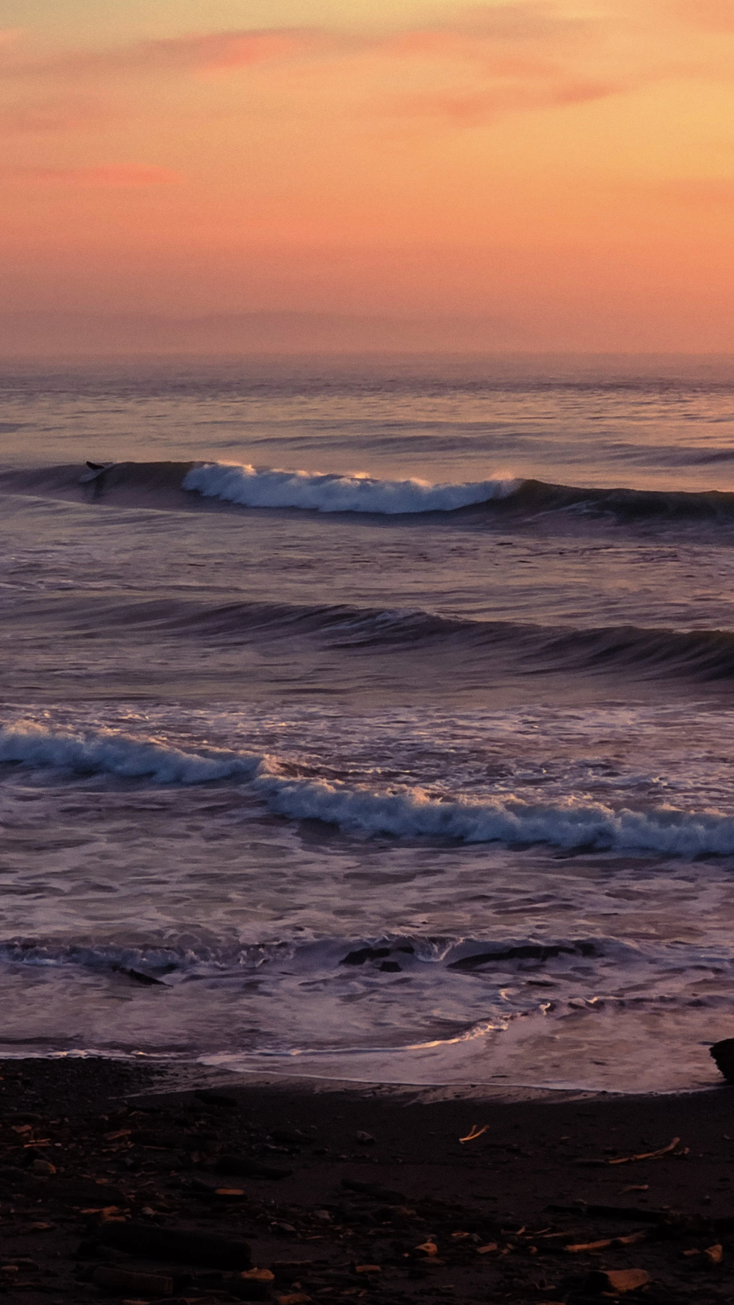 Sunset over the ocean with gentle waves hitting the shore, pink and orange hues in the sky.