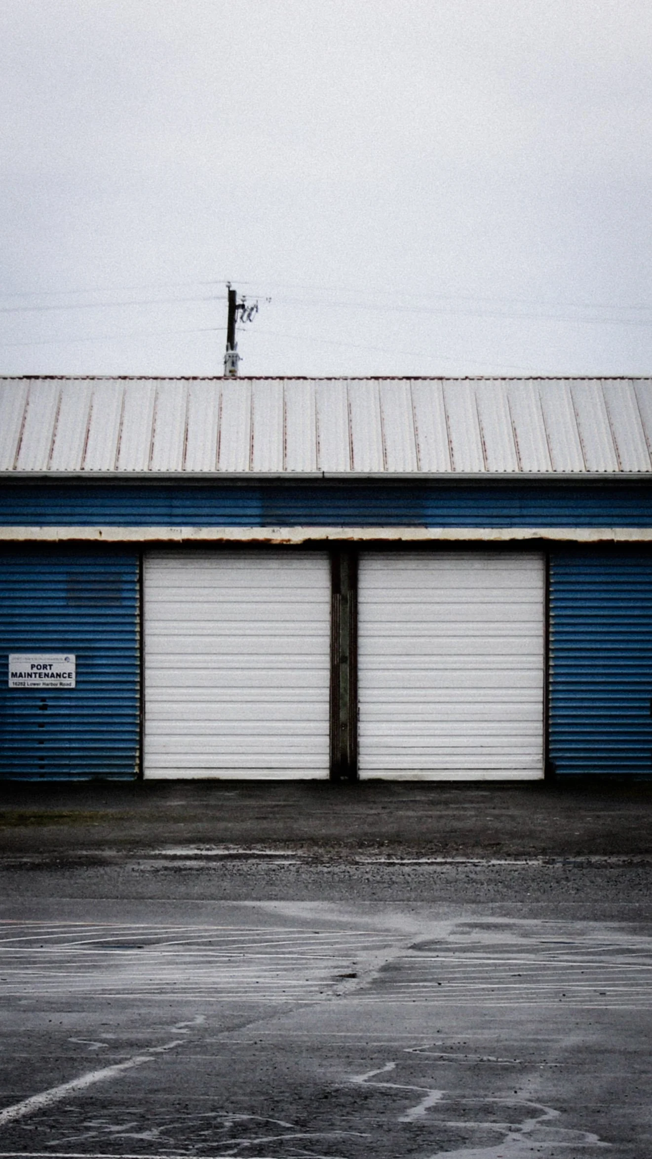 An empty industrial loading dock with a metal gate, a sign reading 'Port Maintenance', and a large roof structure, with an overcast sky.