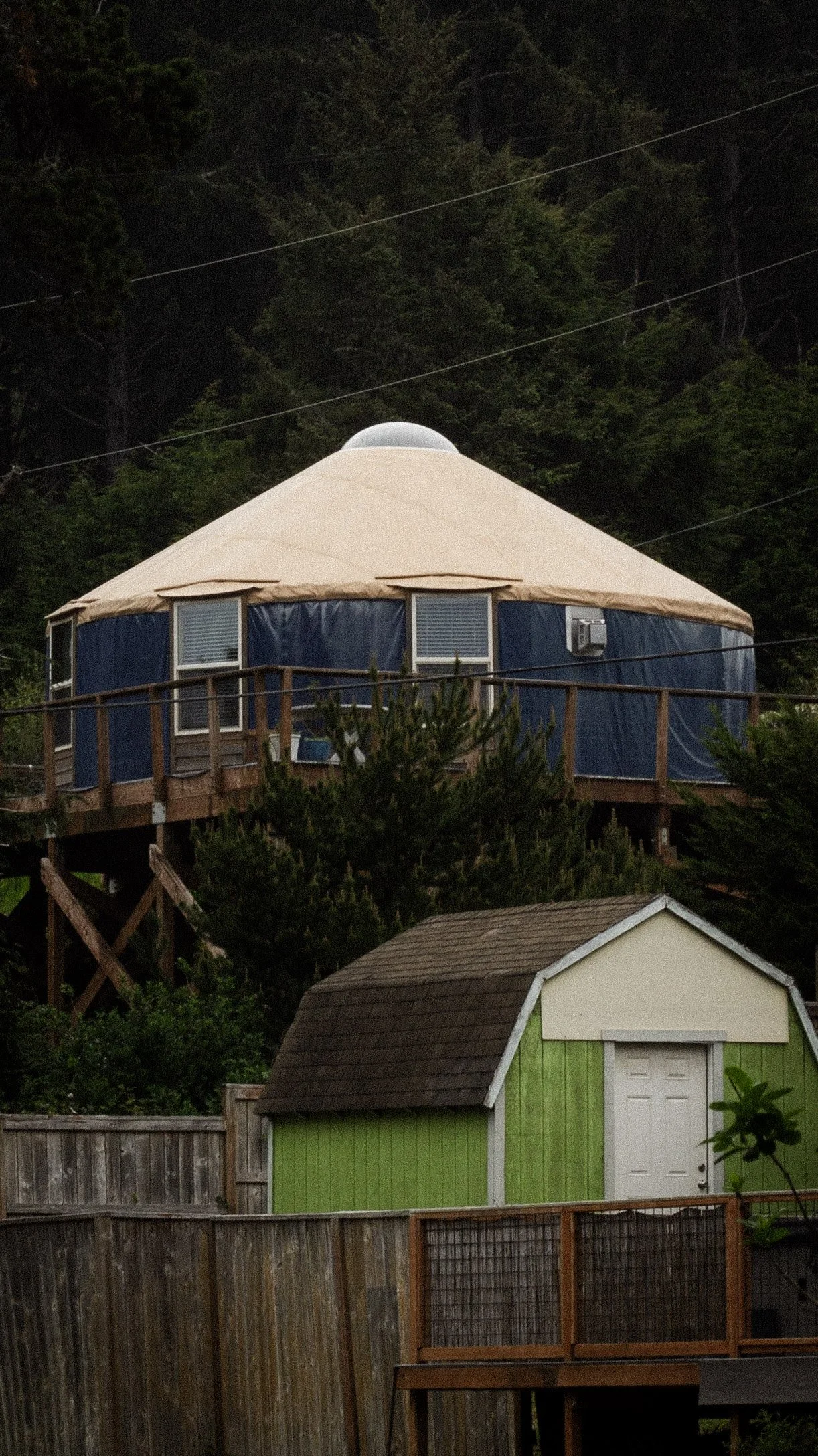 A hilltop yurt with a beige roof, blue sidewalls, and multiple windows, situated among trees with power lines overhead. In the foreground, there are smaller houses or sheds with fences.