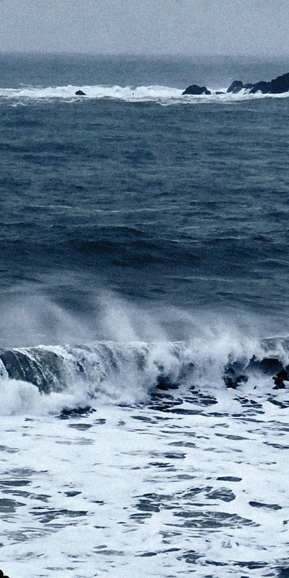 Ocean waves crashing onto rocks with distant waves and overcast sky.