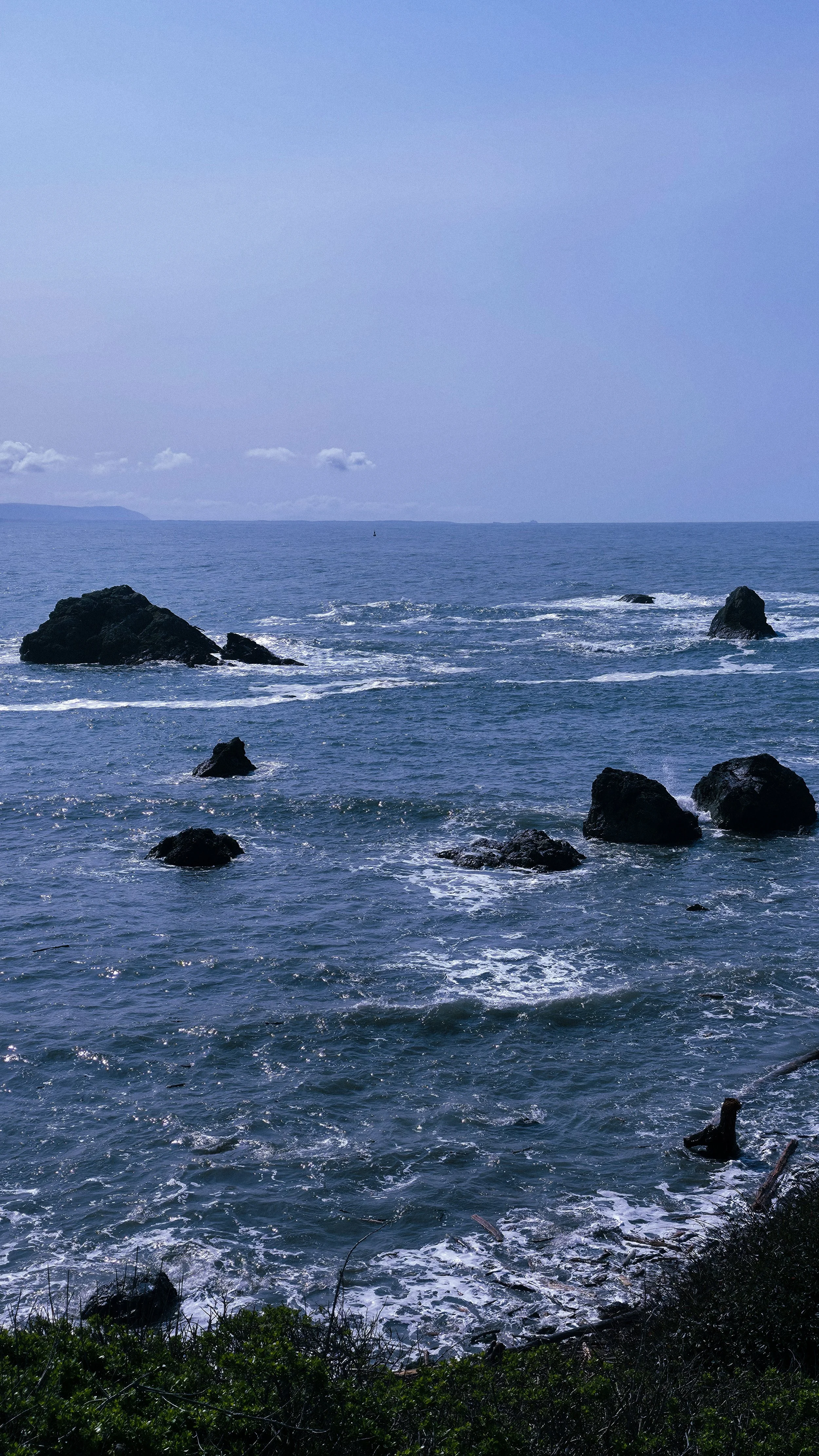 Ocean waves crashing against rocks along the shoreline with a distant island visible on the horizon under a blue sky.