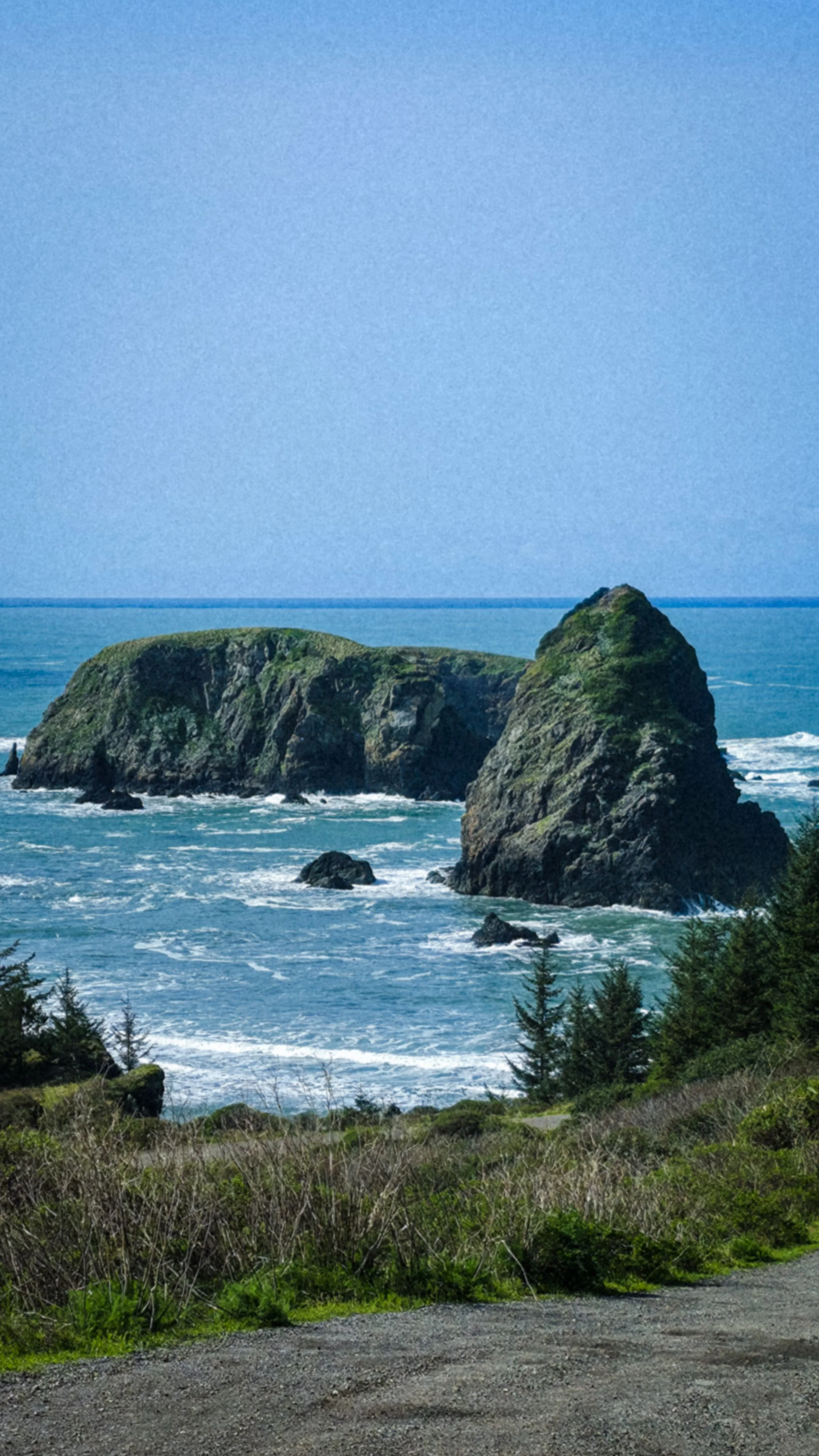 Rocky coastline with ocean waves crashing against large rocks and green hills in the background under a clear blue sky.