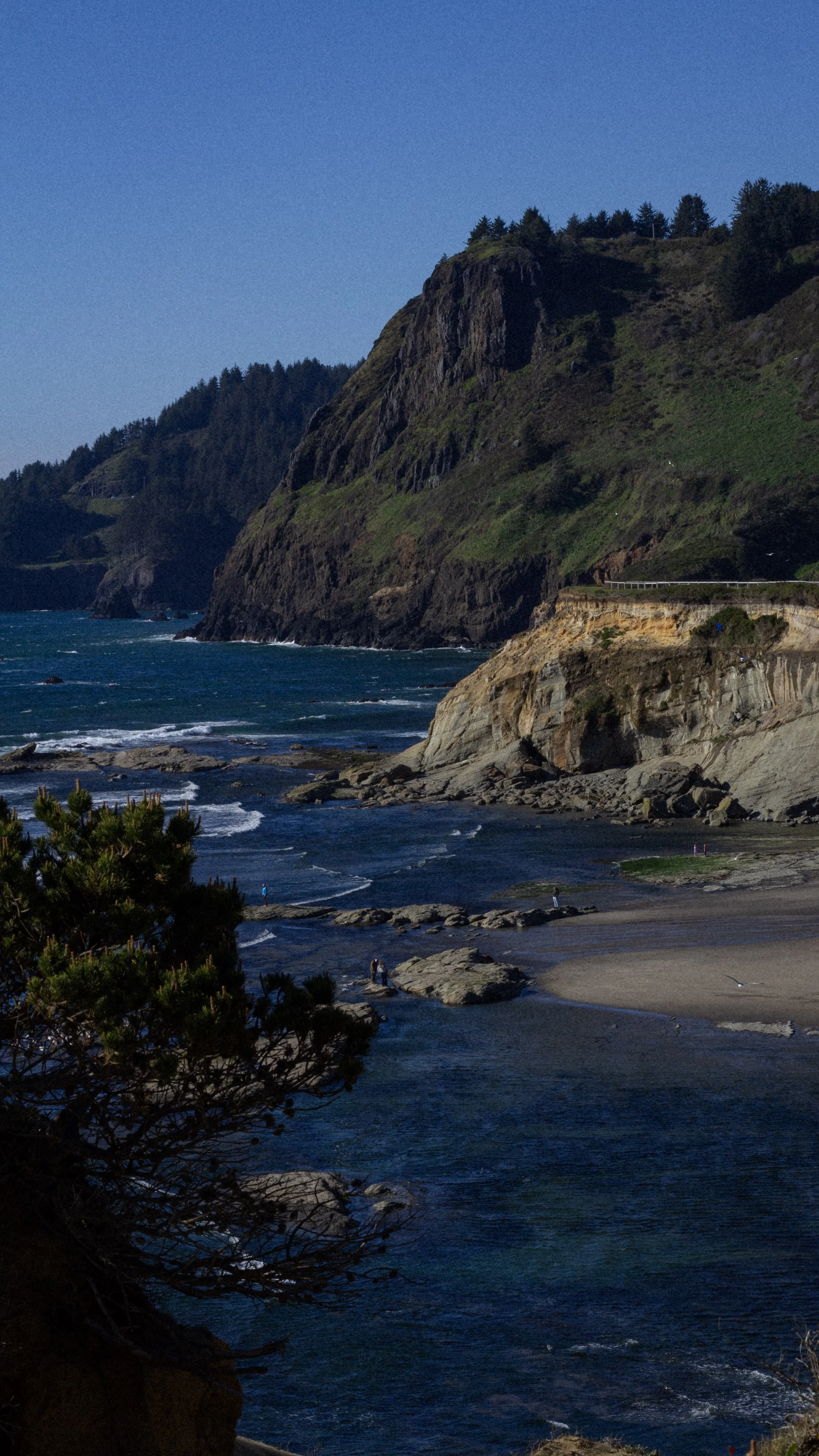 A rugged coastline with steep cliffs covered in greenery, rocky shores, and a few people walking near the water. The ocean waves crash against the rocks, under a clear blue sky.
