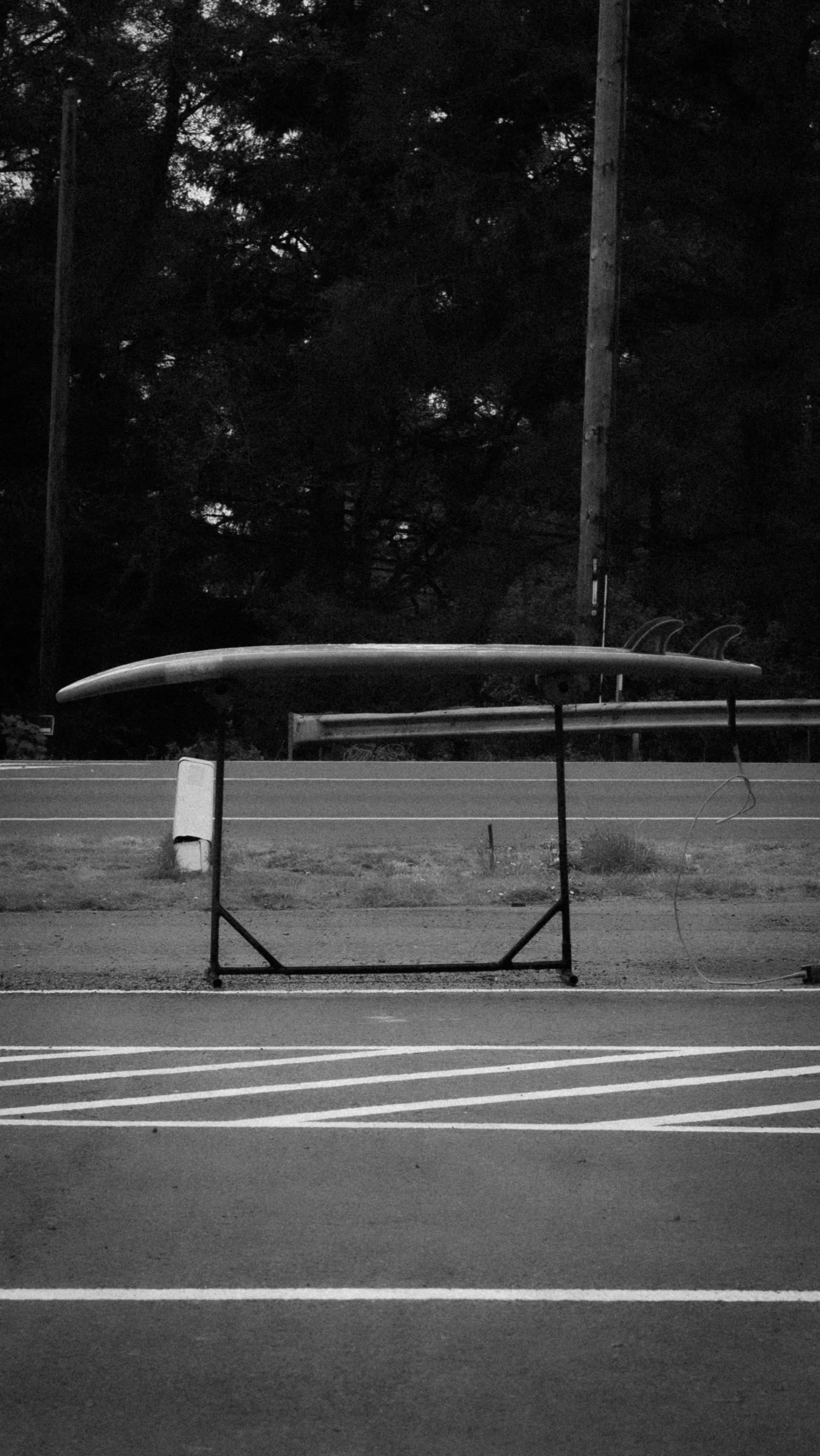 A black and white photograph of a boat on a stand in front of a tree-lined background, with a road or parking lot in the foreground.