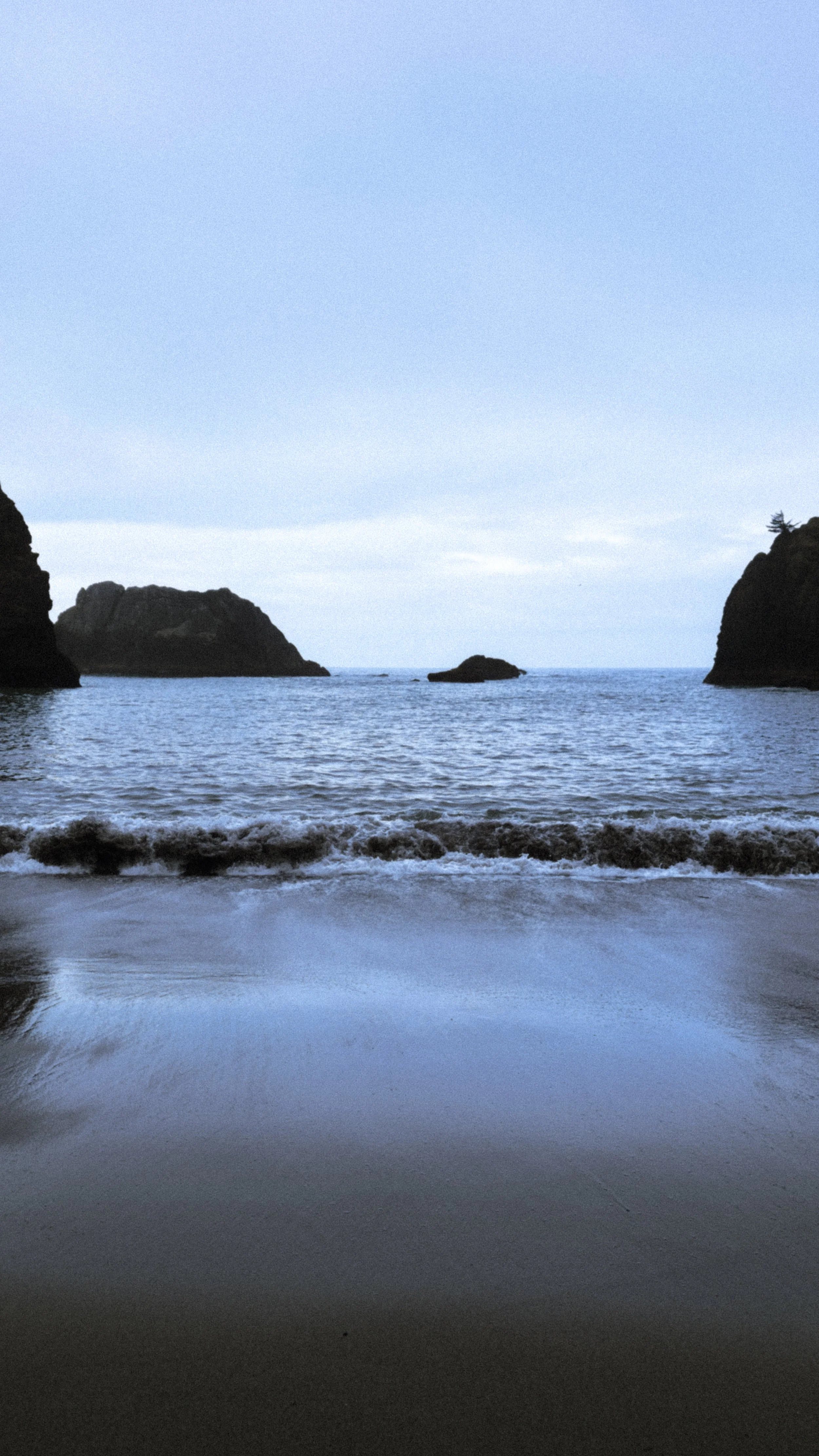 A beach scene with calm ocean water, rocky cliffs on both sides, and small islands in the distance under a cloudy sky.