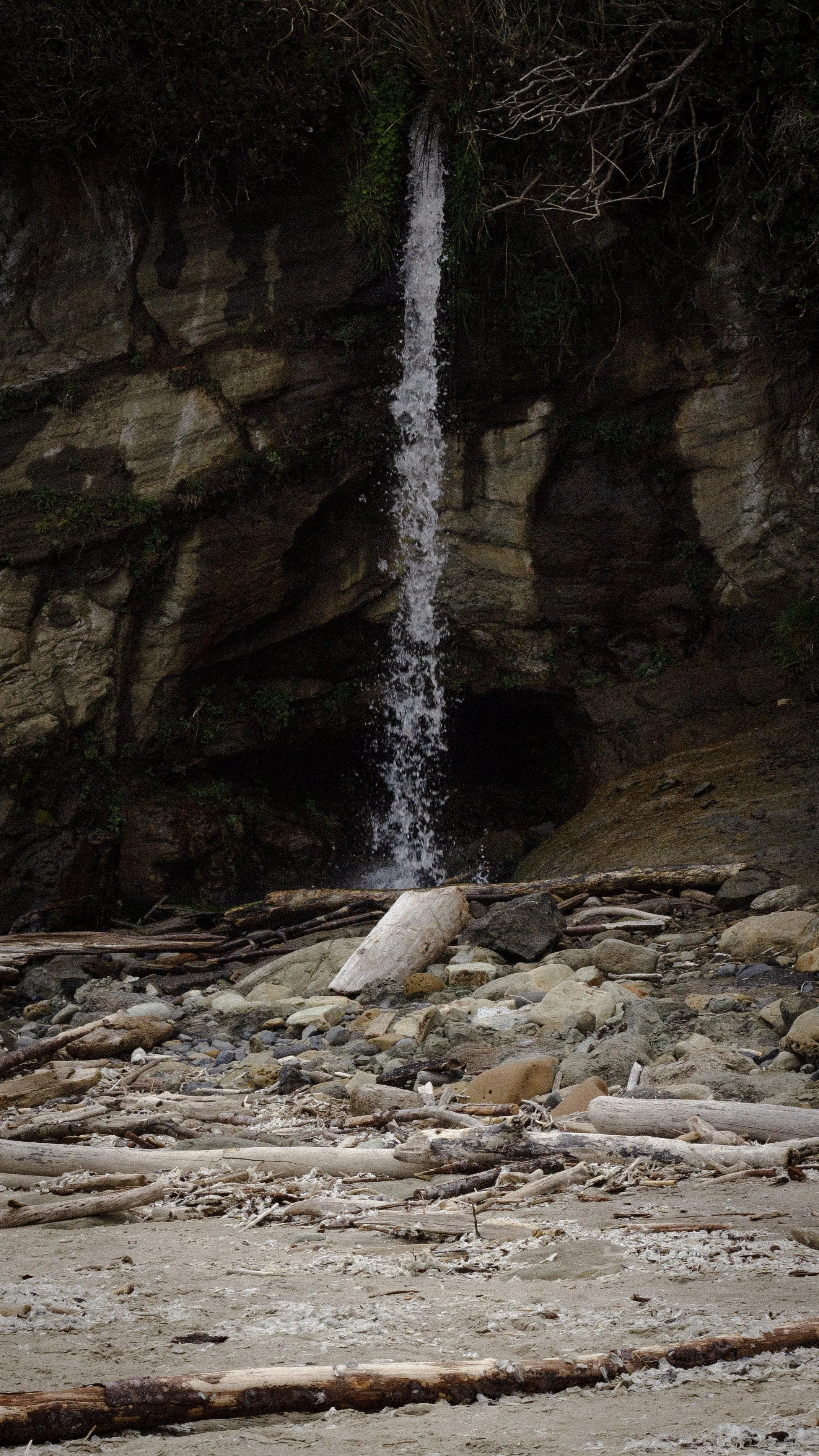 A small waterfall cascading down a rocky cliff surrounded by trees and vegetation, flowing onto a rocky and sandy ground scattered with driftwood and rocks.