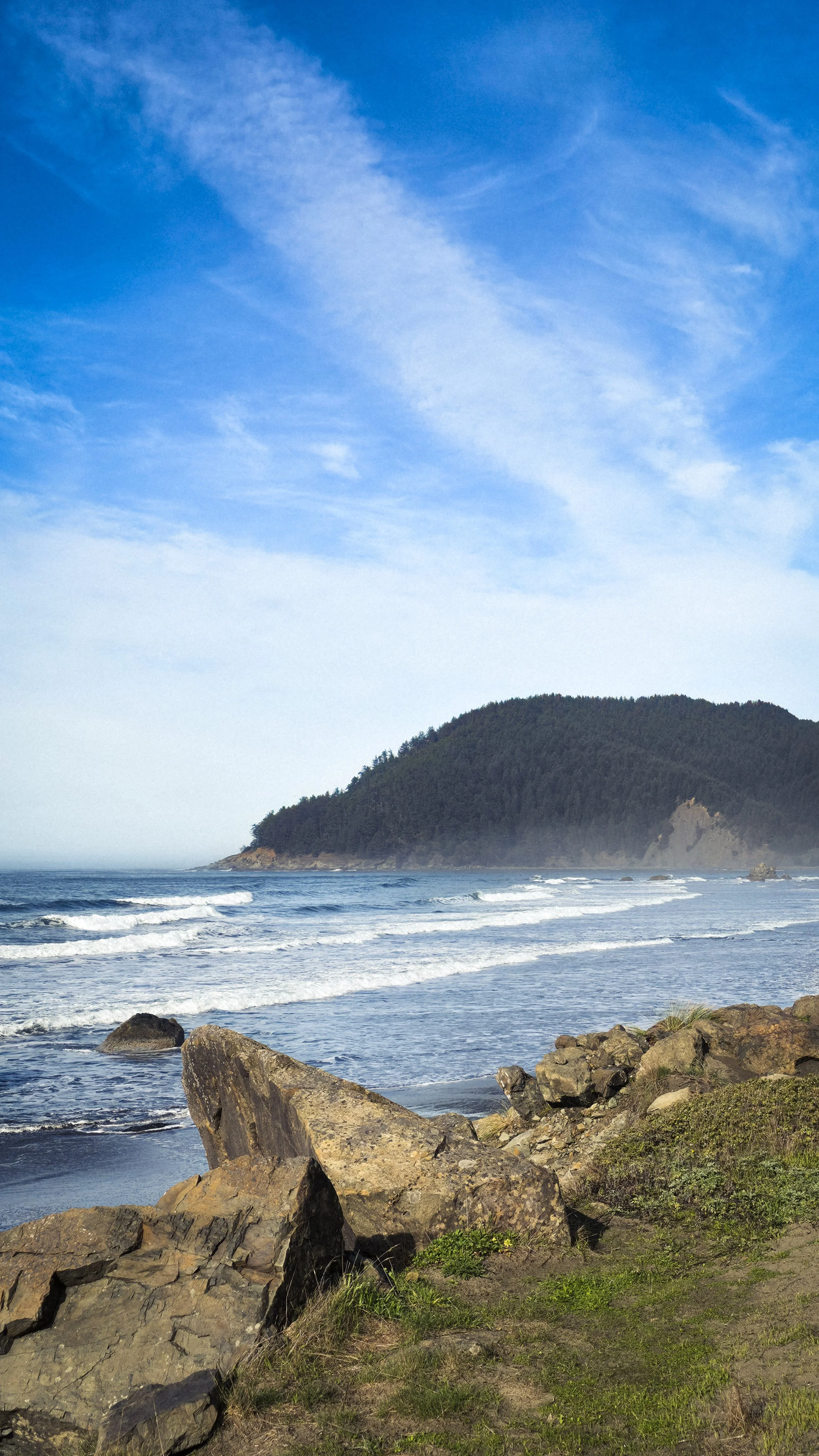 A scenic coastal scene with rocky shoreline in the foreground, waves crashing onto the beach, a forested hill in the middle distance, and a blue sky with wispy clouds above.