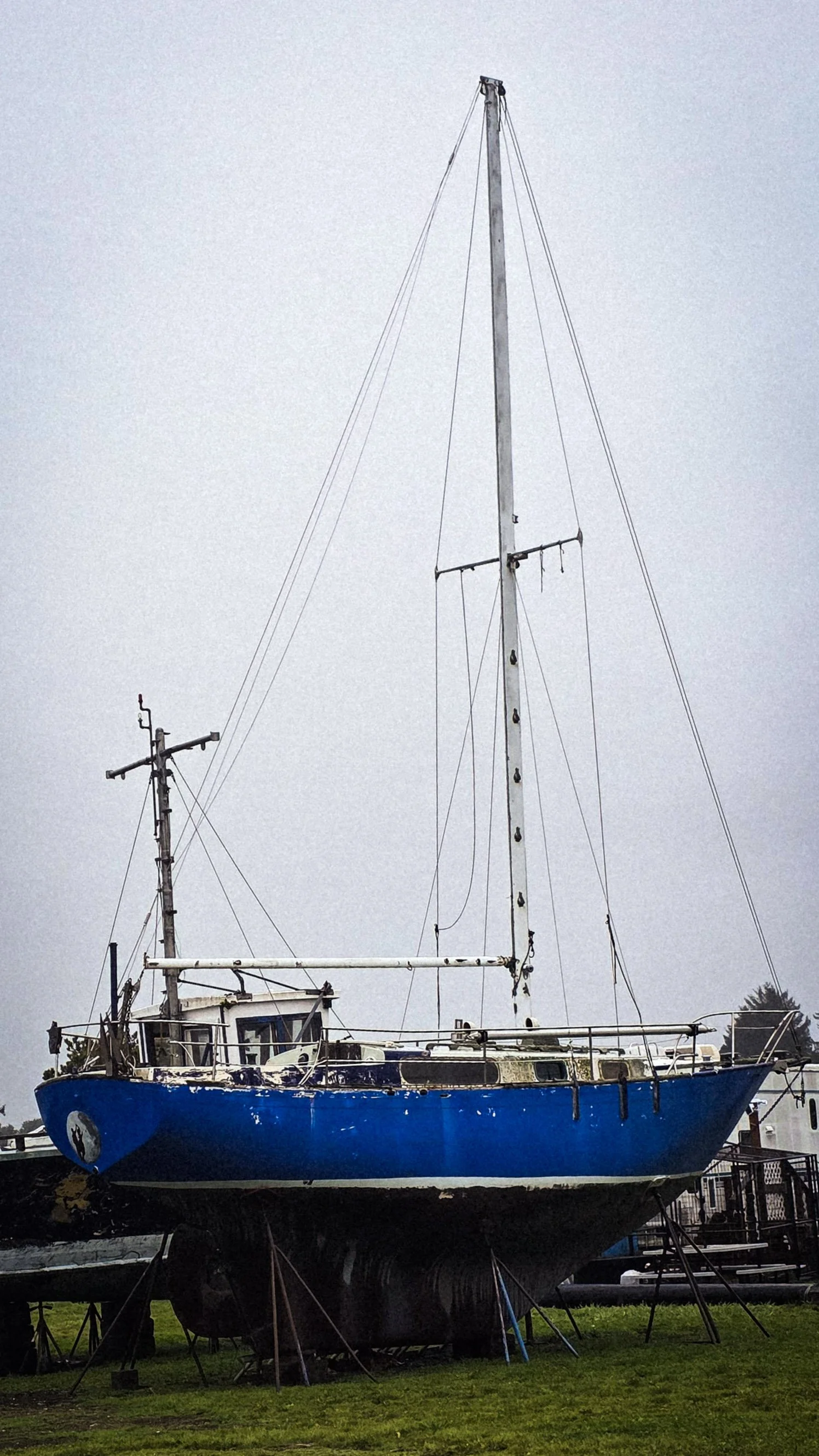 A blue sailboat on land, supported by stands, with a mast and rigging against a gray sky.
